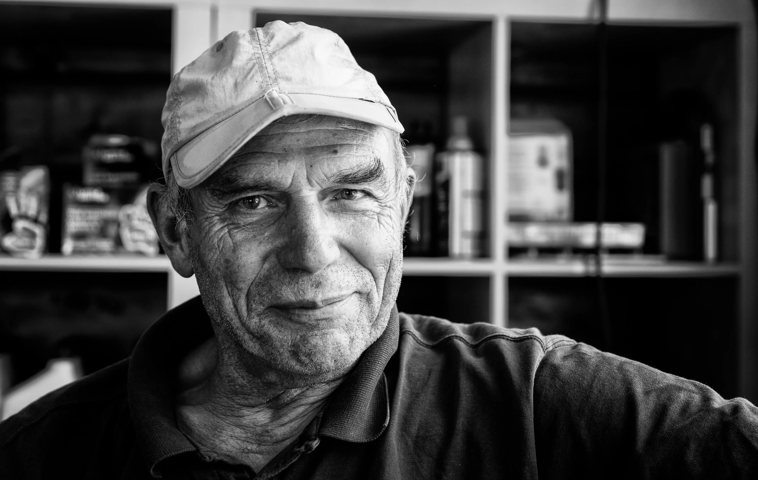 A black and white photo of an elderly man with a cap slightly tilted back, showing a confident expression in front of a background with shelves and boxes.