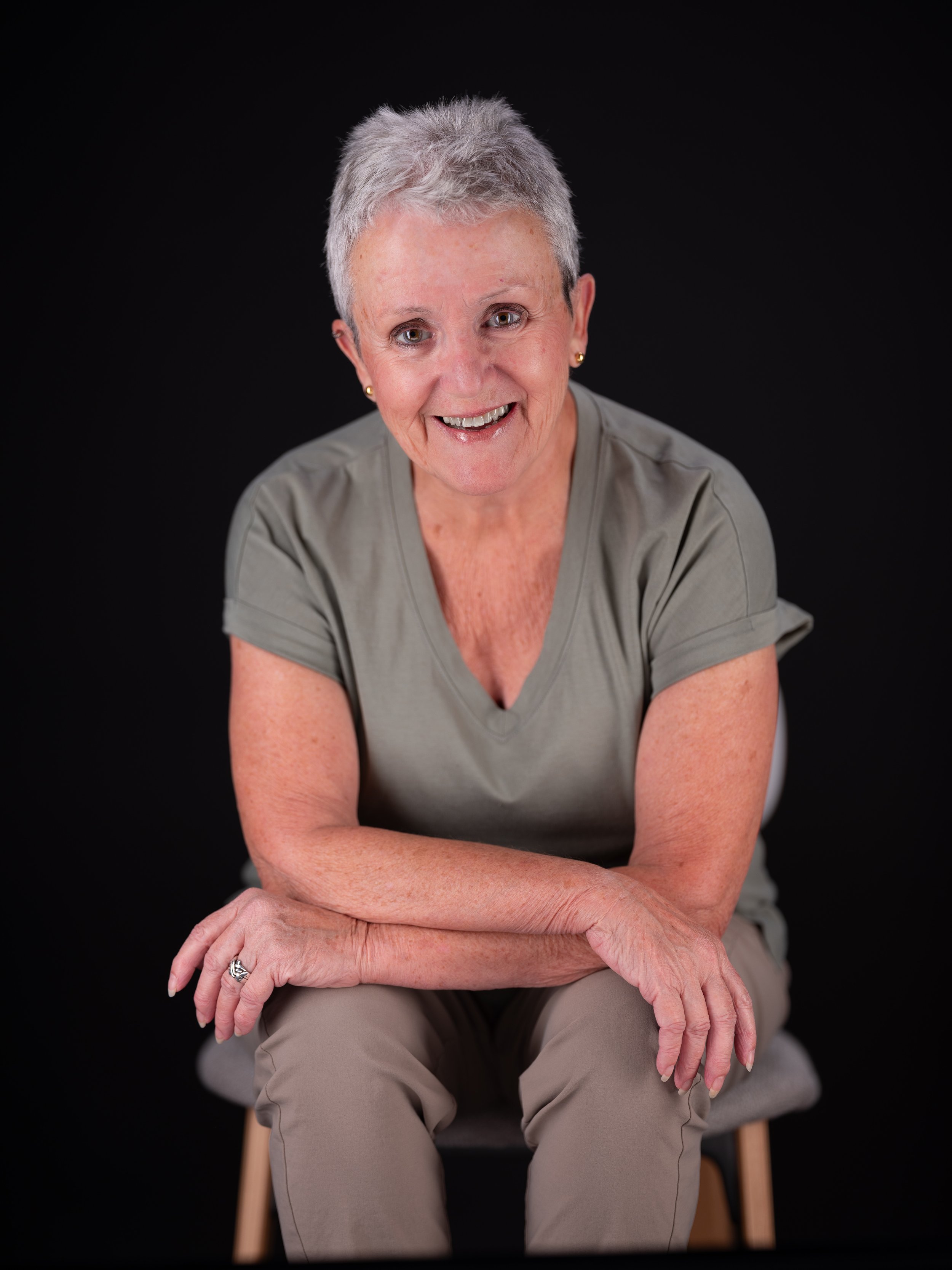 Older woman with short gray hair smiling, sitting on a wooden chair against a black background, wearing a light gray T-shirt and khaki pants.
