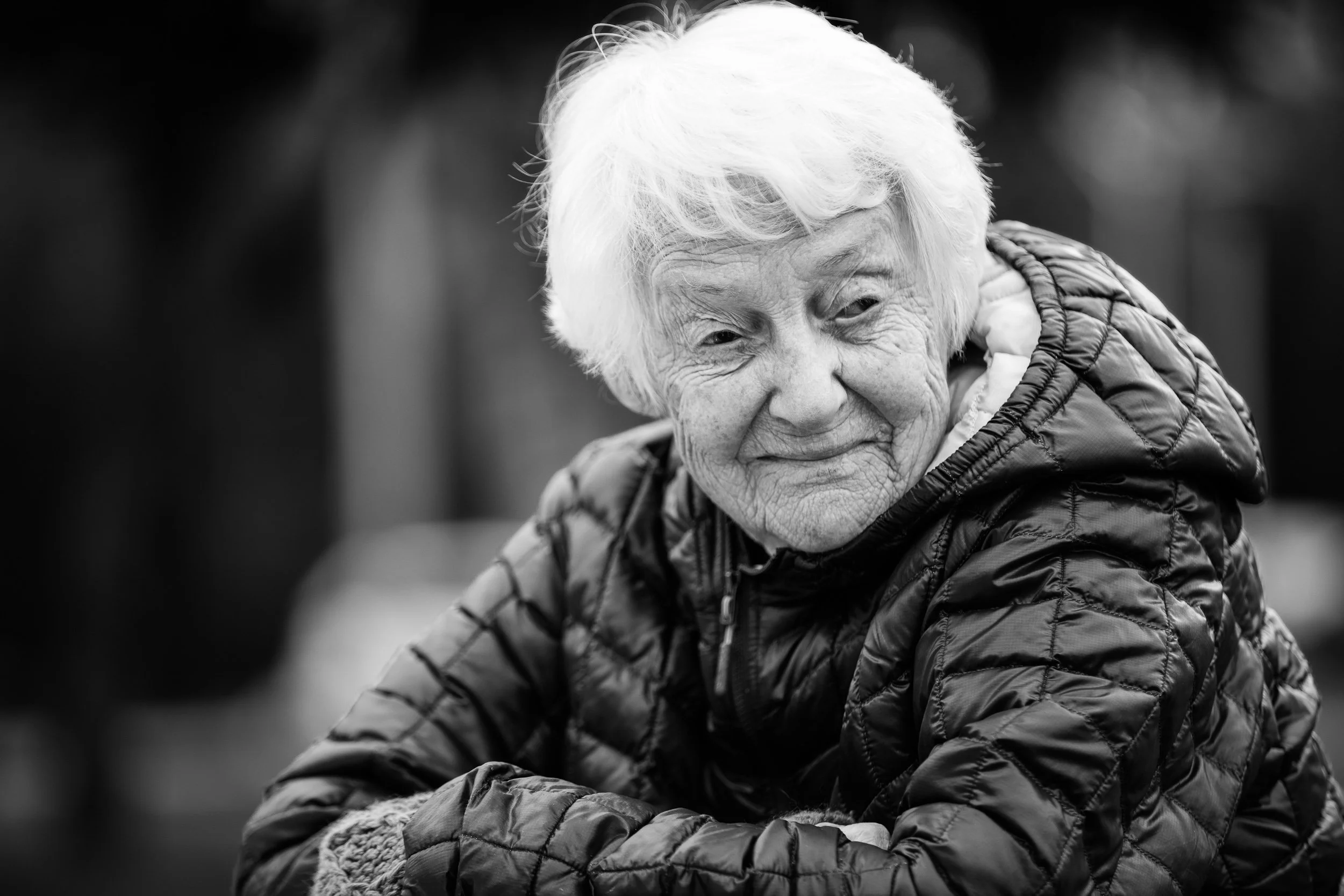 A black and white photo of an elderly woman smiling, wearing a quilted jacket, with her head tilted slightly downward and her eyes looking to the side.