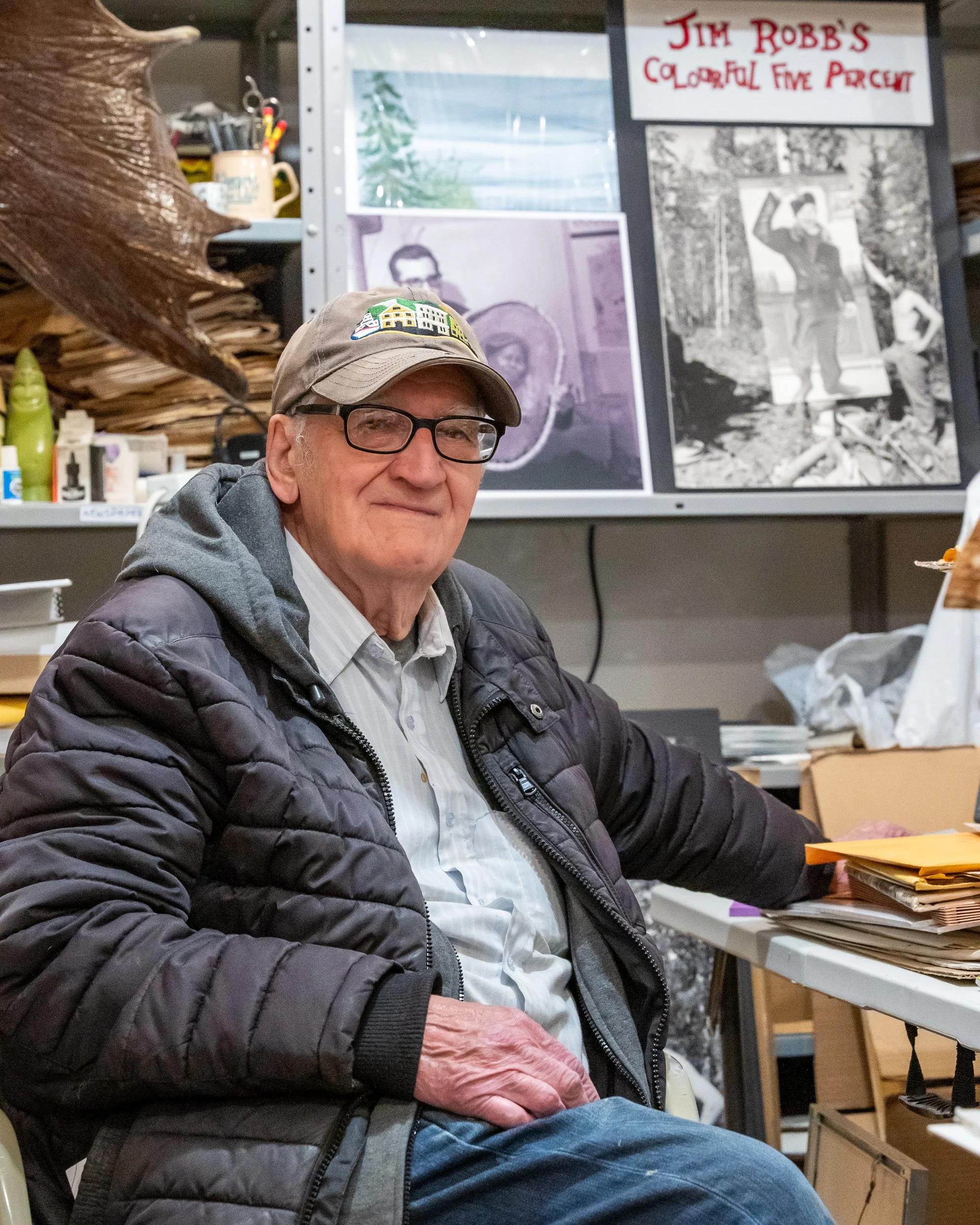 A man wearing glasses, a beige cap with a colorful pattern, a white shirt, a gray hoodie, and a black quilted jacket is sitting at a desk. Behind him is a display with black-and-white photos and a sign that reads 'Jim Robb's Colorful Five Percent.' T