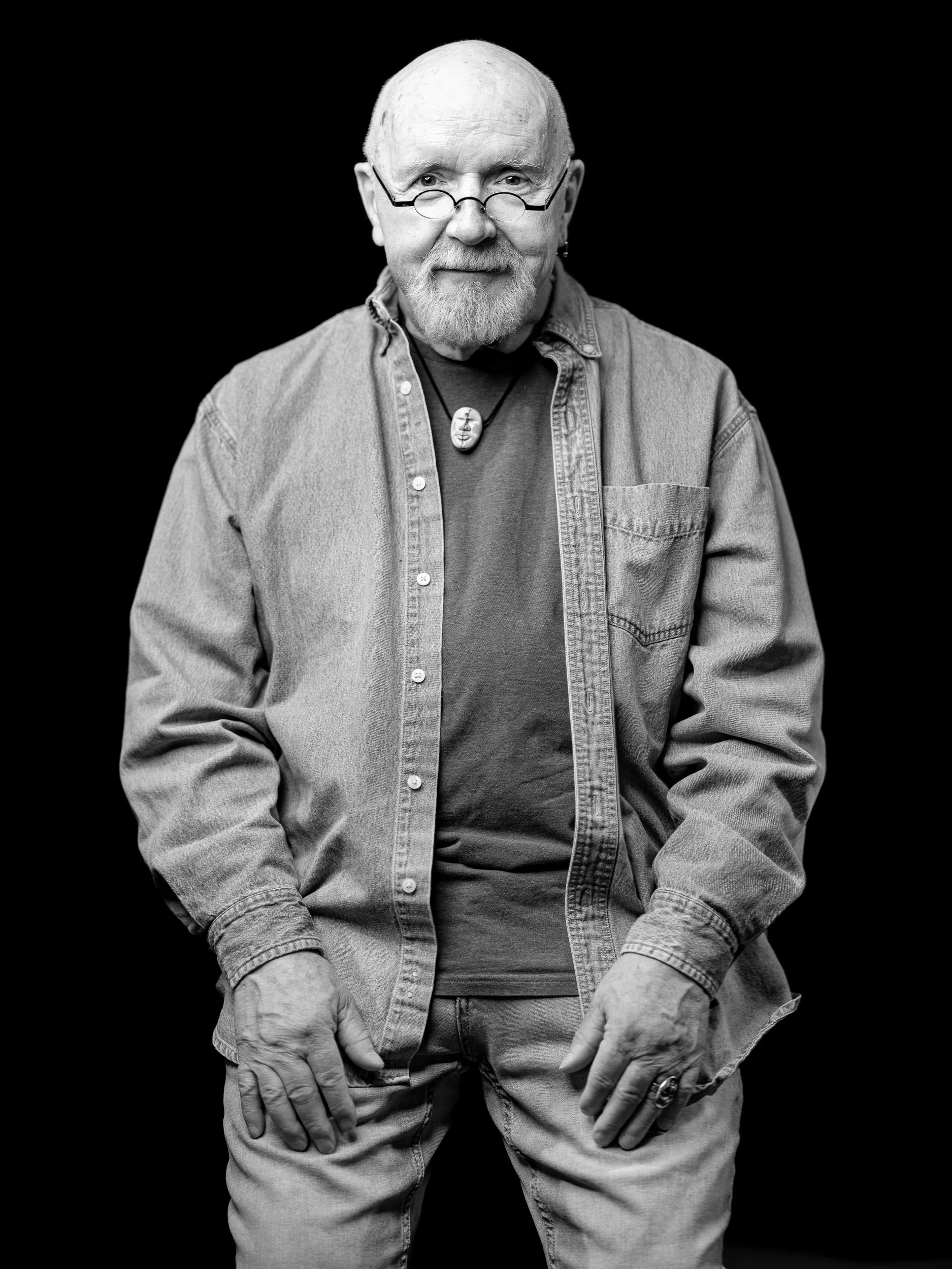 Black and white portrait of an elderly man with a beard, glasses, and a earring, wearing a denim shirt and a necklace with a face-shaped pendant, posing against a black background.