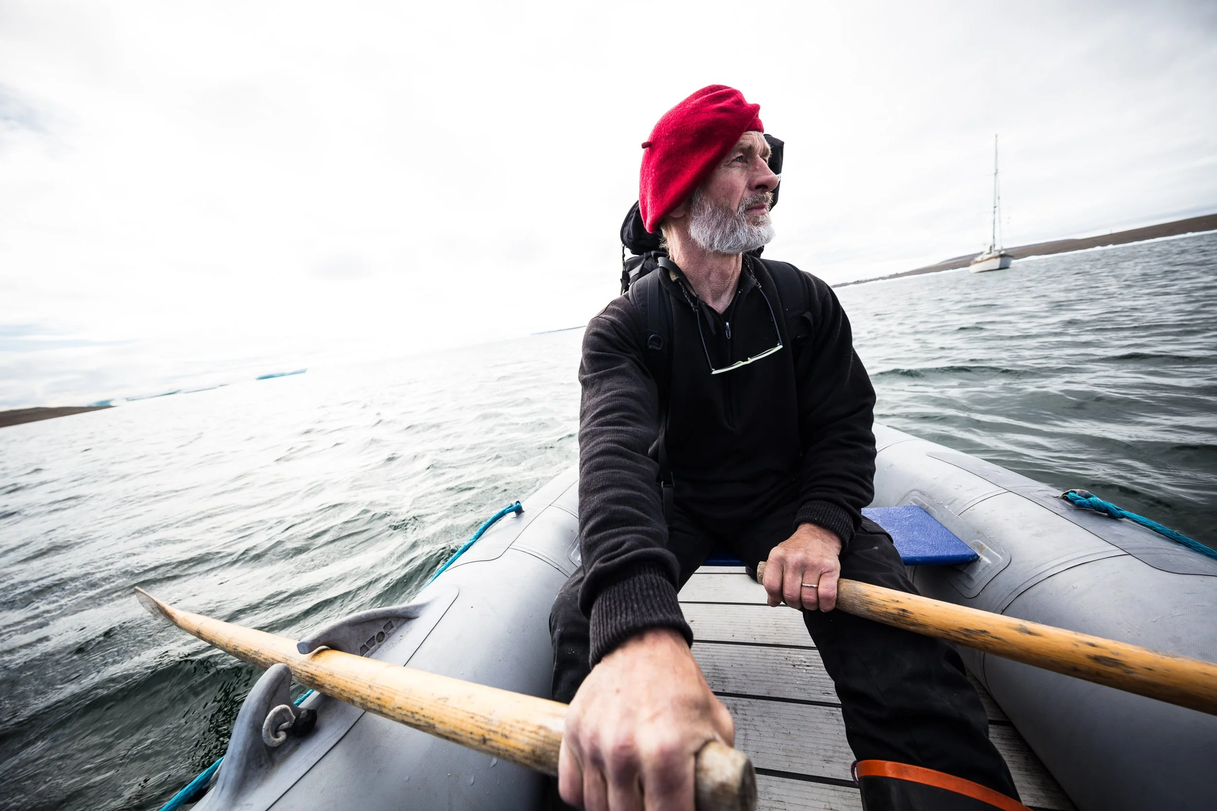 An older man with a gray beard wearing a red beanie and black jacket paddles a small boat on water, with a sailboat in the background under a cloudy sky.