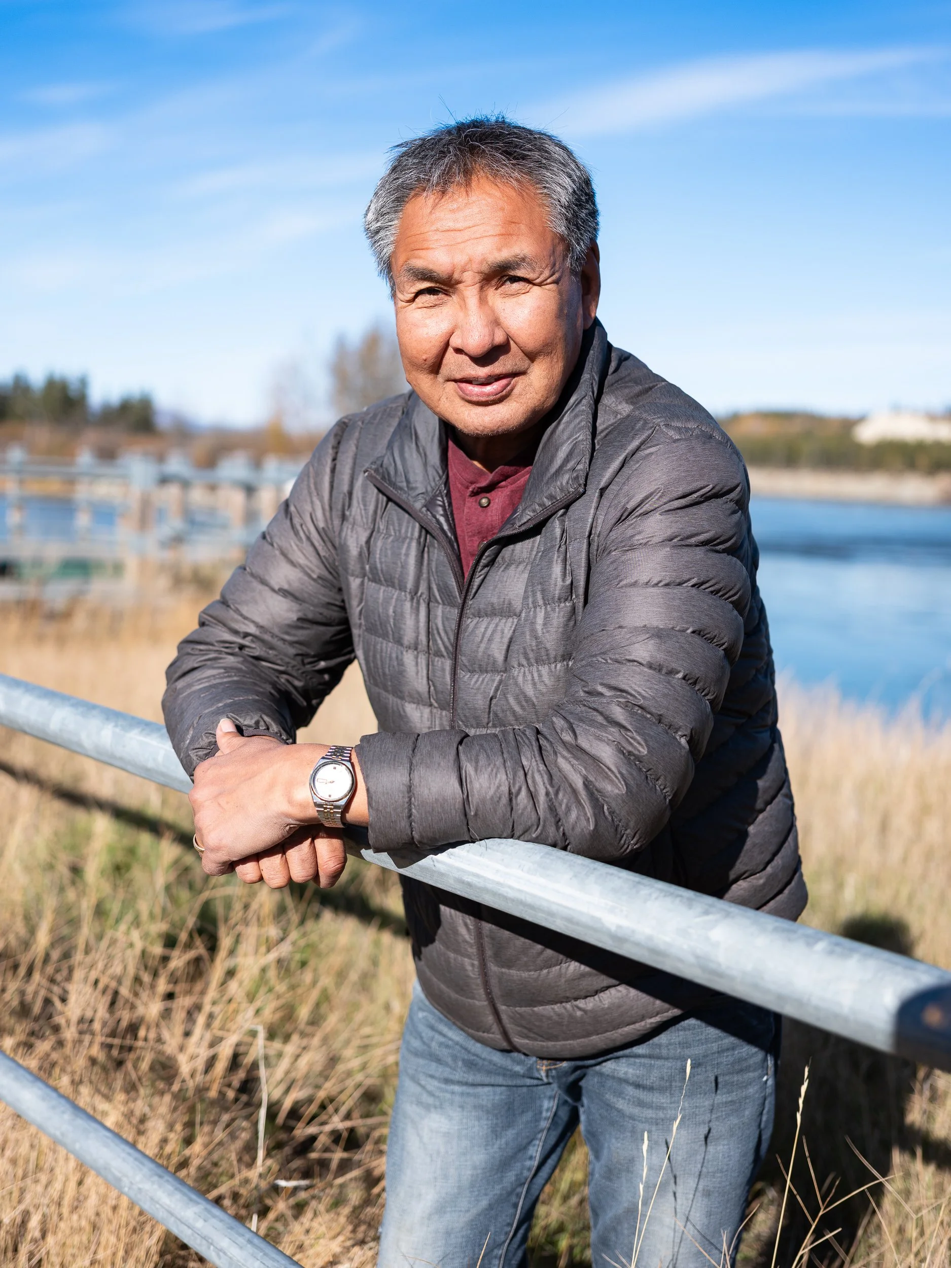 A middle-aged man with gray hair and a slight smile, wearing a gray puffy jacket, a maroon shirt, and a silver watch, leaning on a metal railing outdoors during daytime with a body of water and trees in the background.