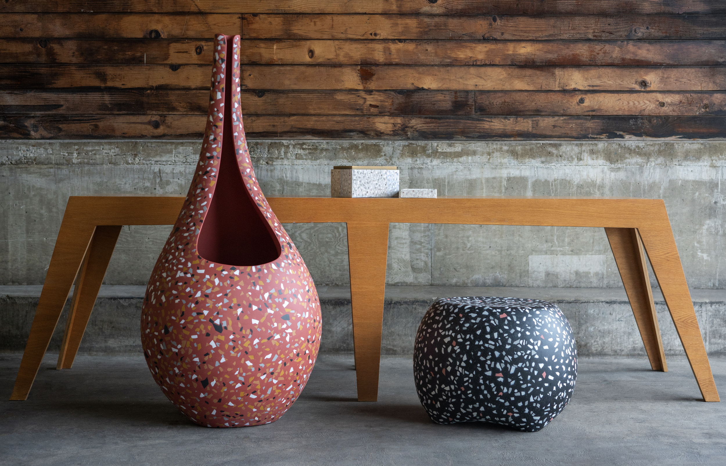 Modern furniture set featuring a red, speckled ceramic sculpture, a wooden angular table, and a black, patterned ottoman in an industrial setting.