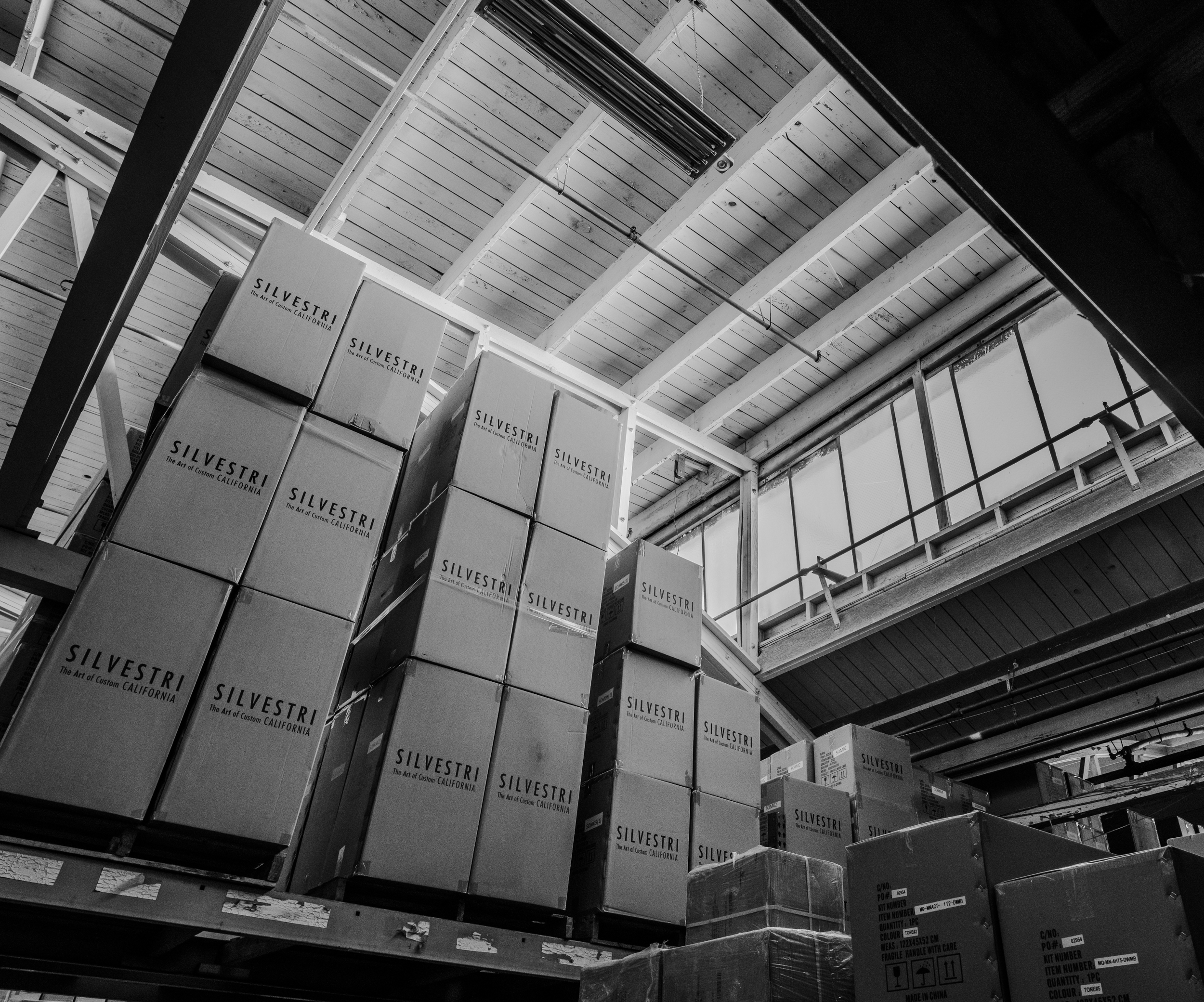 Black and white photo of stacked cardboard boxes labeled 'Silvestri' in a warehouse with high ceilings and large windows.