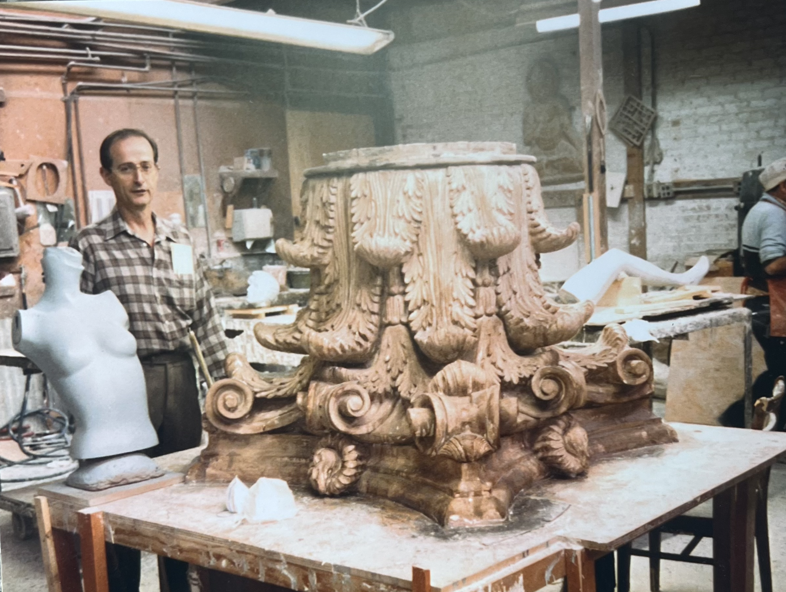 Man in workshop with ornate sculpture, mannequin bust, and various tools and materials, demonstrating craftsmanship and artistry.
