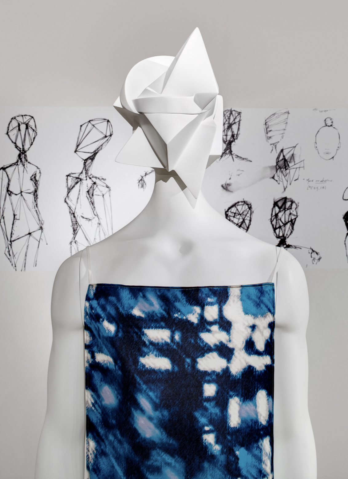 Sculpture with an angular white headpiece and blue patterned clothing, with sketched figures in the background.
