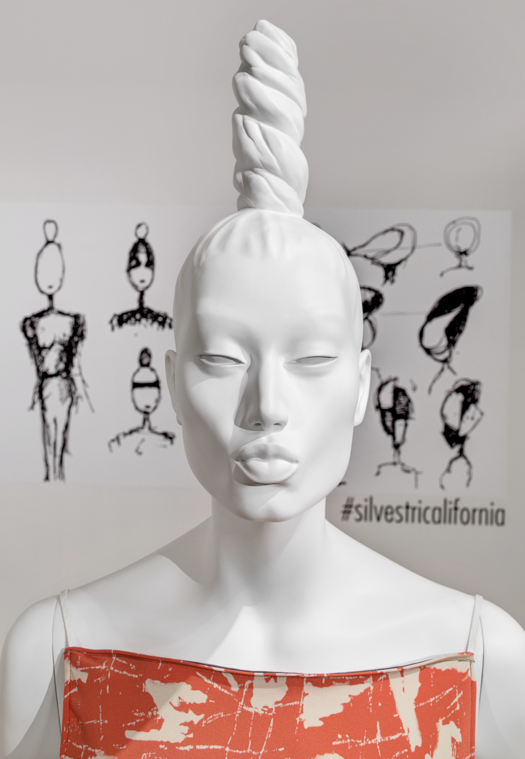 White mannequin with twisted hair design, wearing a red patterned top, in front of abstract fashion sketches on a wall.