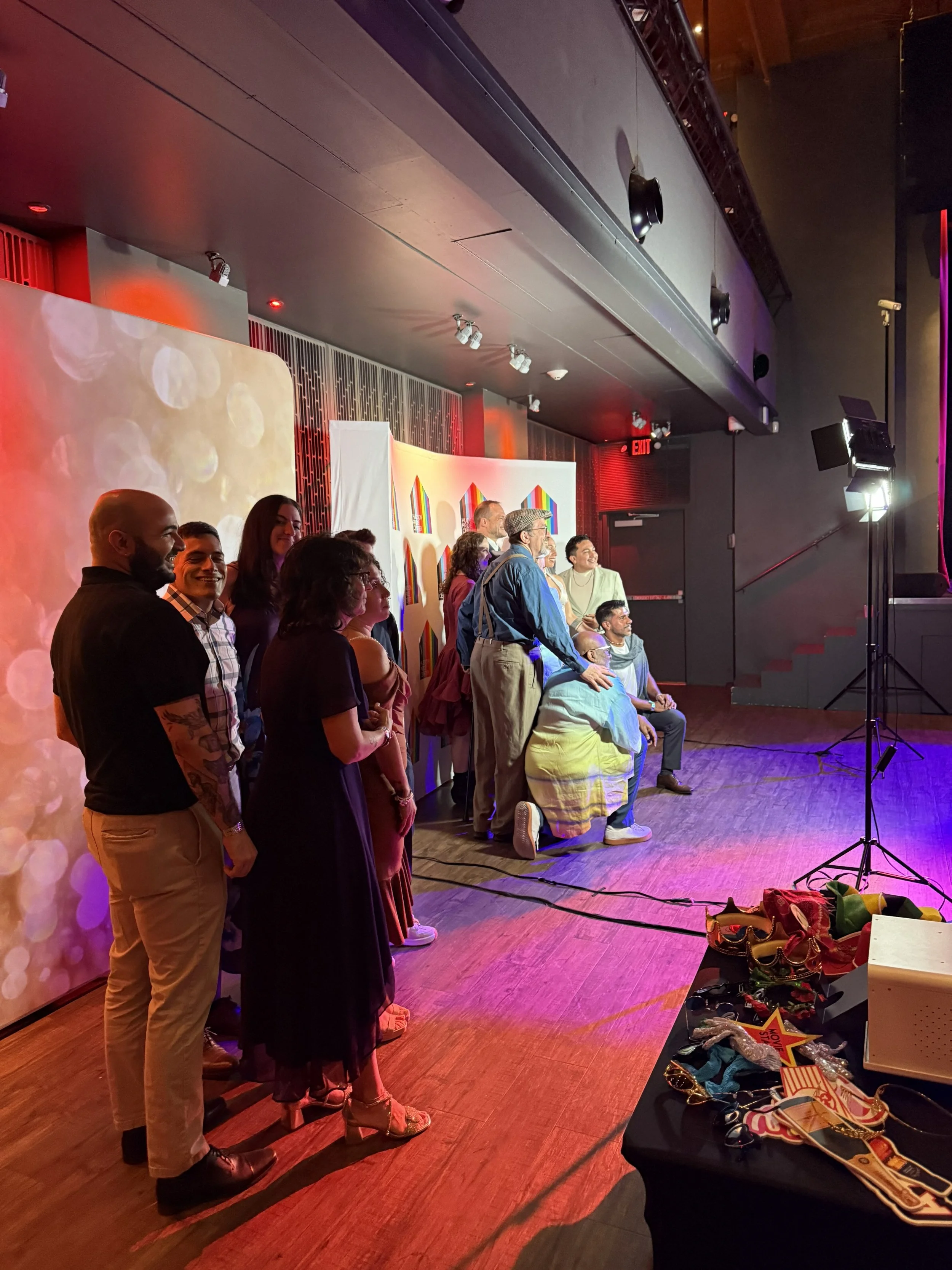 A group of people standing in a line on a stage. They are facing to the right. The stage has a colorful backdrop with rainbow-colored arches. There are items placed on a table in the foreground, including jewelry, pins, and bags. The room is lit with