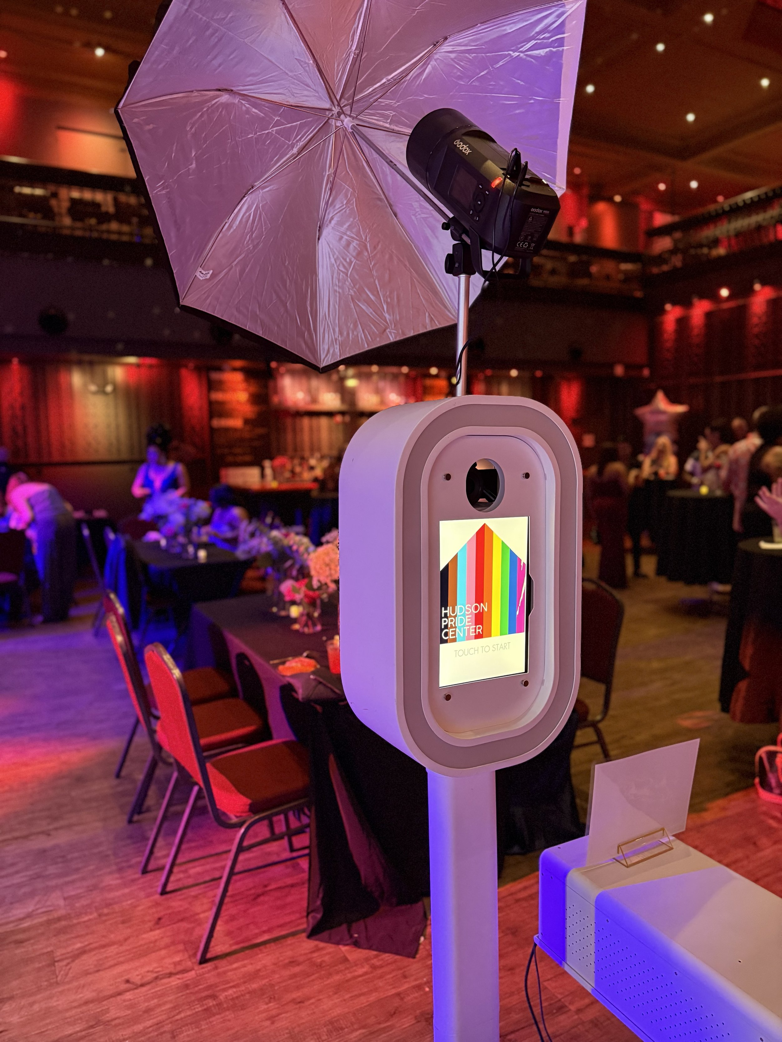 Photo of a ticket kiosk at the Hudson Pride Center event, featuring a purple umbrella attached to the top, with people and tables in the background under dim lighting.