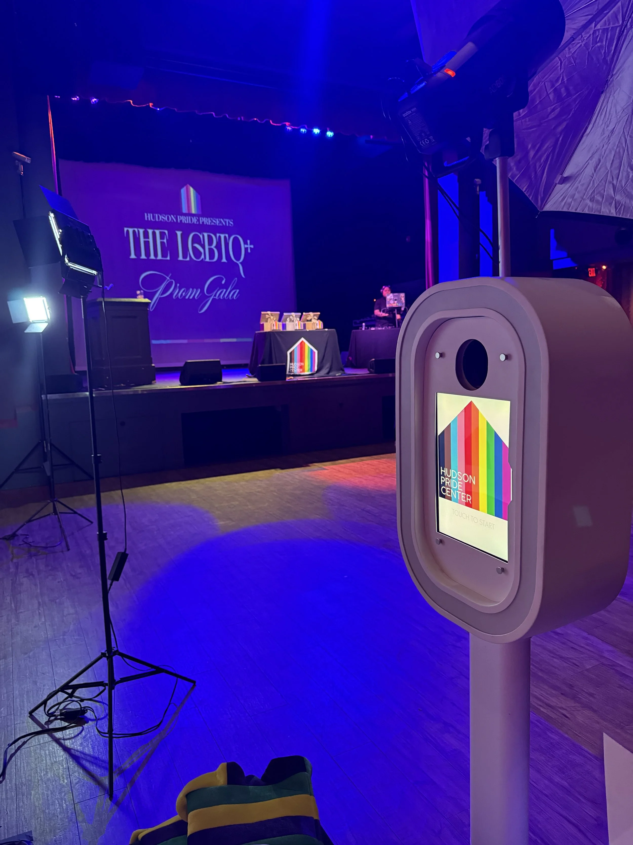 Stage with a large screen displaying event title, tables with gifts, DJ or sound technician, and colorful lighting at an indoor event for the LGBTQ+ community.