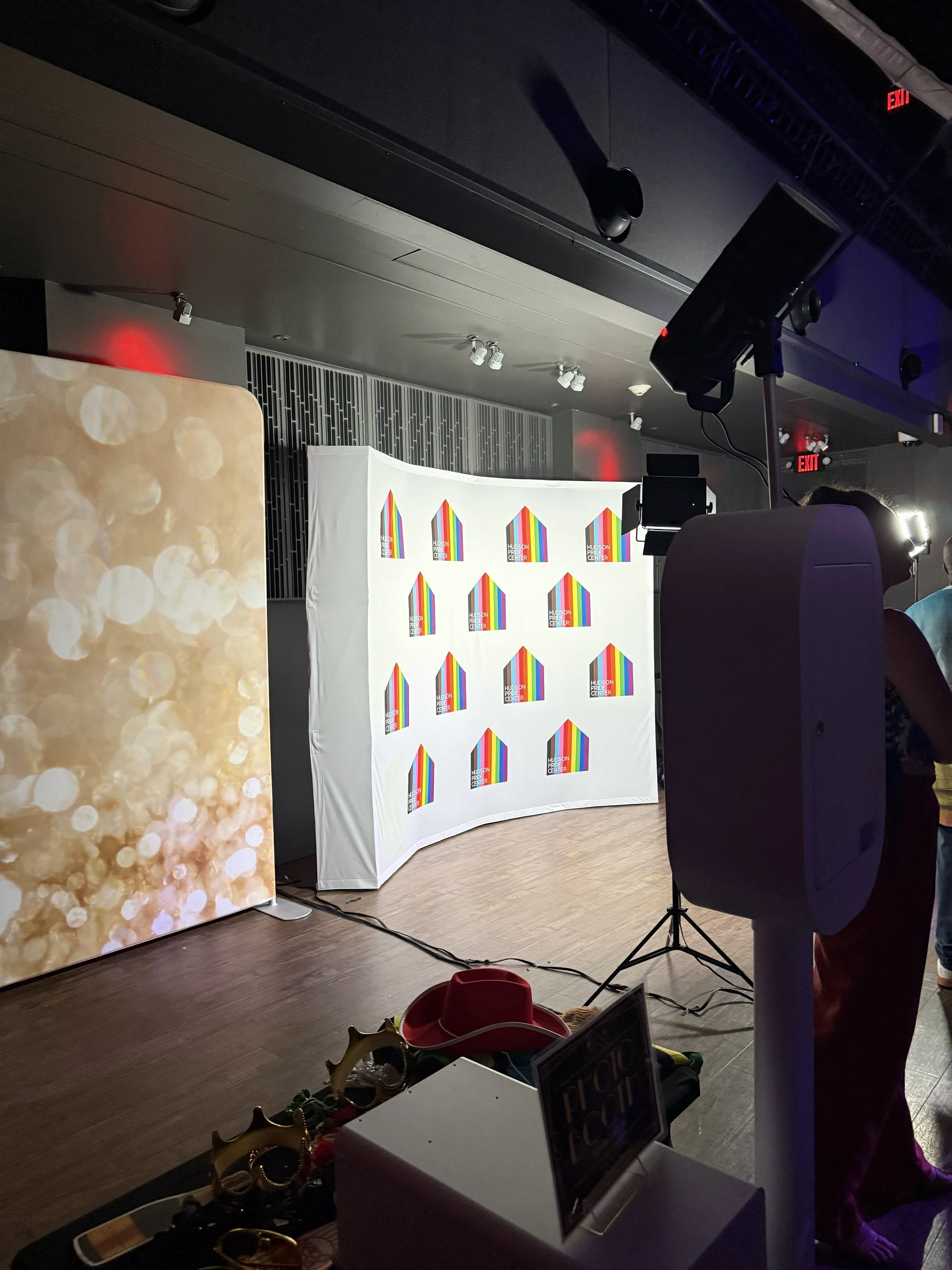 A photo of a stage setup at an event space with a backdrop featuring rainbow-colored stripes and text that reads 'HUDSON PRIDE CENTER.' There are overhead lights, a speaker in the foreground, and decorative holiday props on a table at the bottom of t