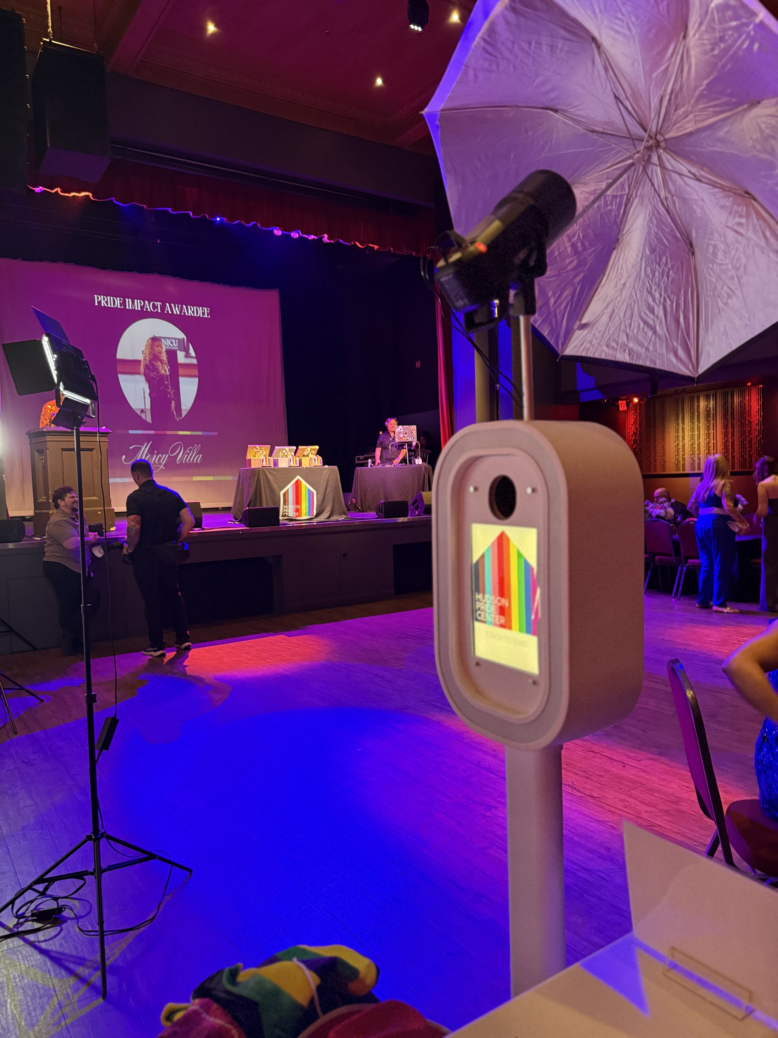 A stage with a large screen displaying 'PRIDE IMPACT AWARDEE' and a photo of a woman named Alicia Villa, with some awards on a table. There is equipment like lights and a microphone in the scene. Audience members are visible, and a large umbrella-sha