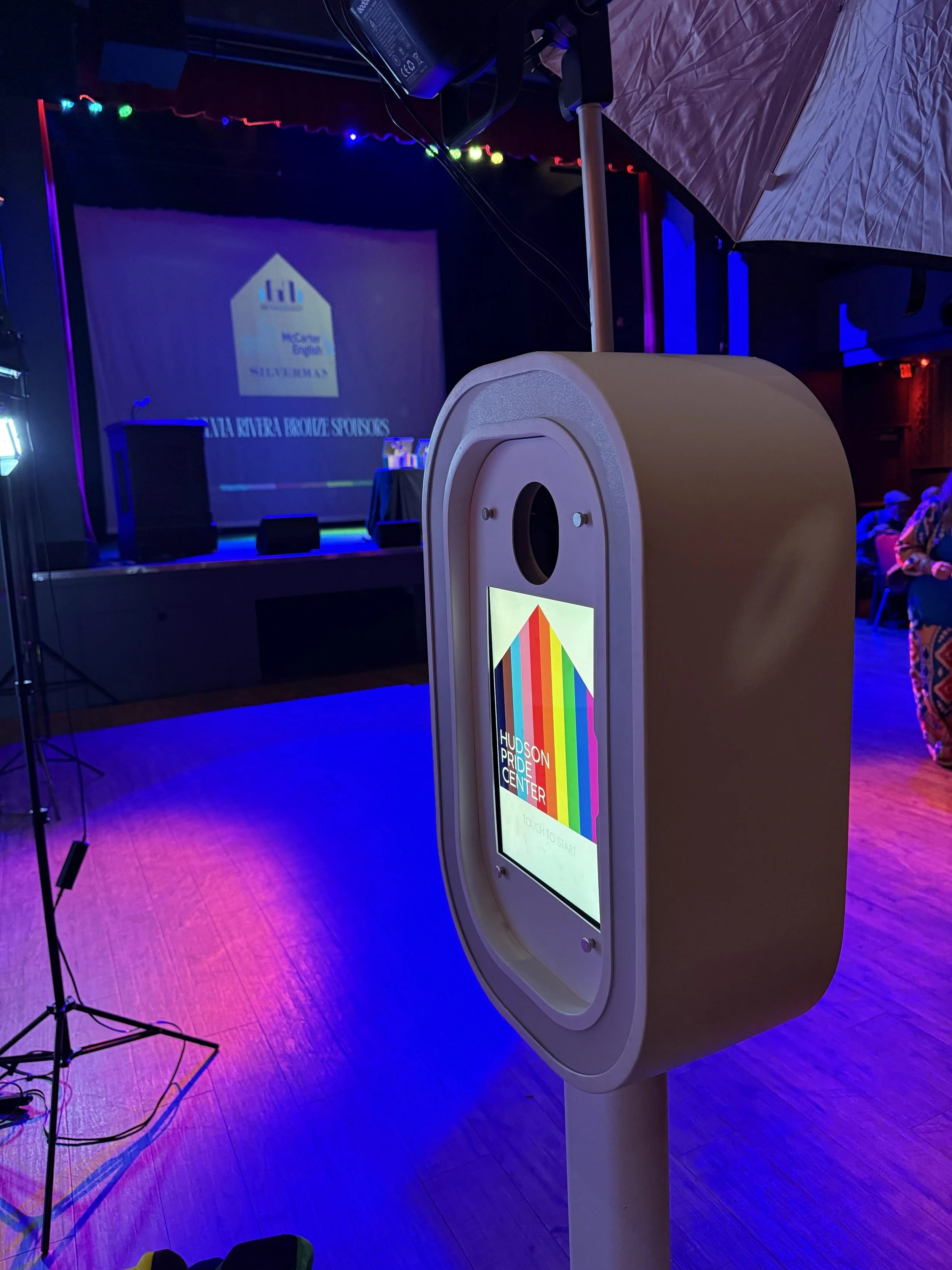 A photo of a voting or check-in kiosk at a Pride event, with a colorful rainbow warning and text that reads "Hudson Pride Center". In the background, a stage with a screen displaying the Hudson River Bravura Sponsors logo and some audience members se