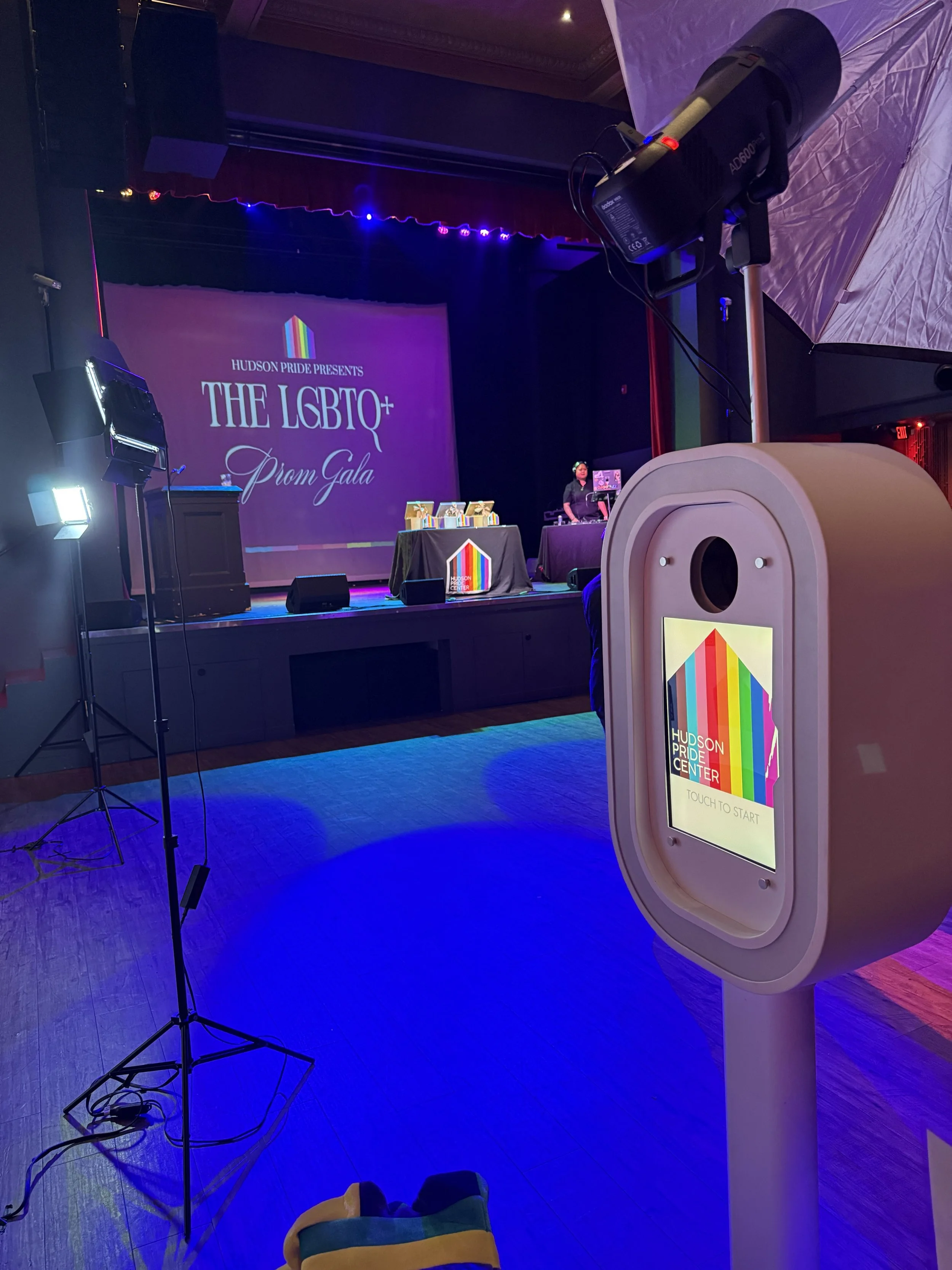 Stage with large screen displaying 'Hudson Pride Presents The LGBTQ+ Prom Gala' with rainbow-colored logo, DJ or performer standing at a table with equipment, and various lights and equipment in the venue.