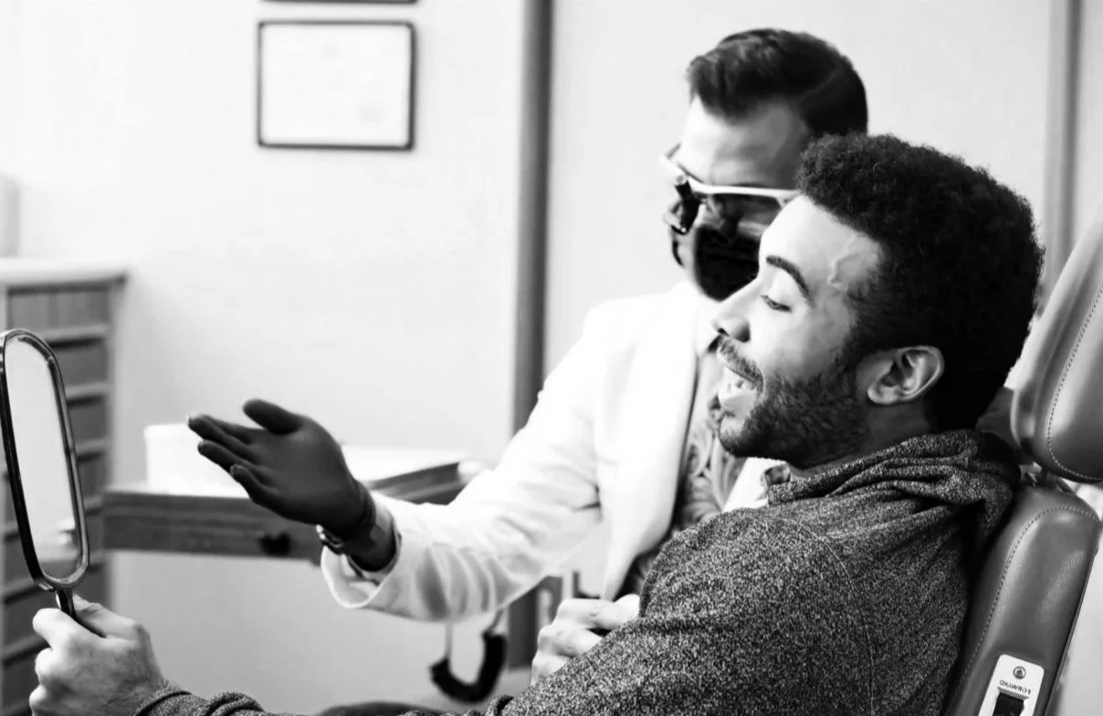 A black man sitting in a dental chair, smiling, while a dental professional wearing gloves, a mask, and protective glasses shows him something in a mirror.