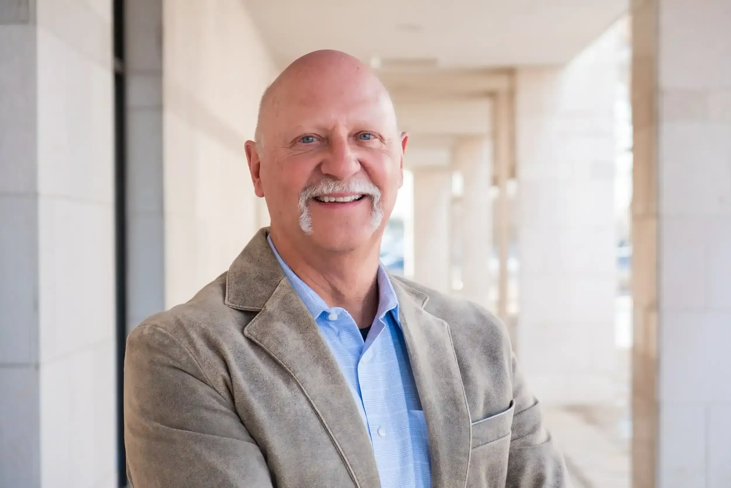 A smiling older man with a mustache and goatee, wearing a light brown blazer and a blue shirt, standing in a well-lit corridor.