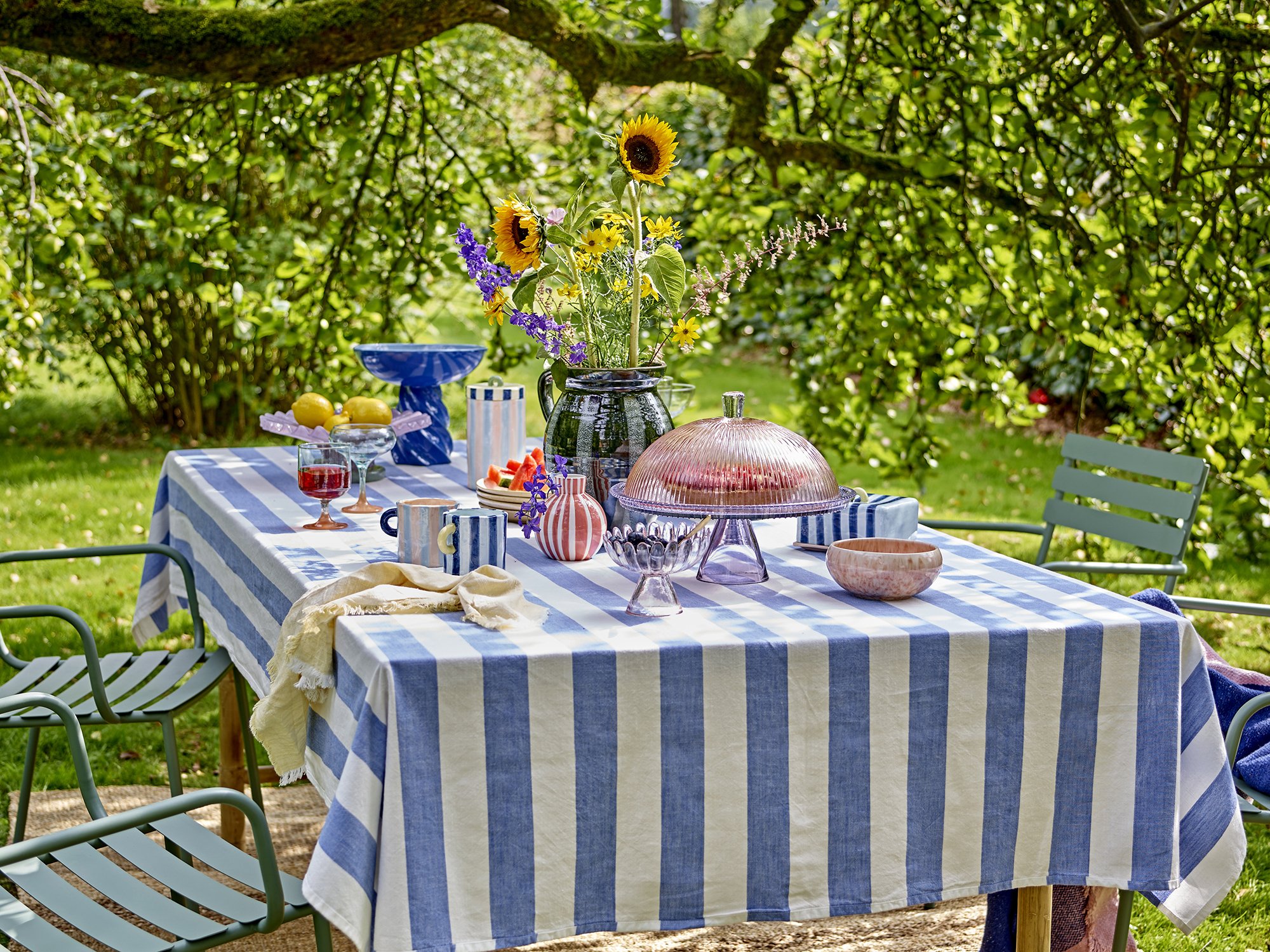 Mesa decorada con un mantel de rayas azules y blancas, con jarrones de flores, frutas y vajilla de colores de la marca Bloomingville, en un jardín rodeado de árboles y arbustos.