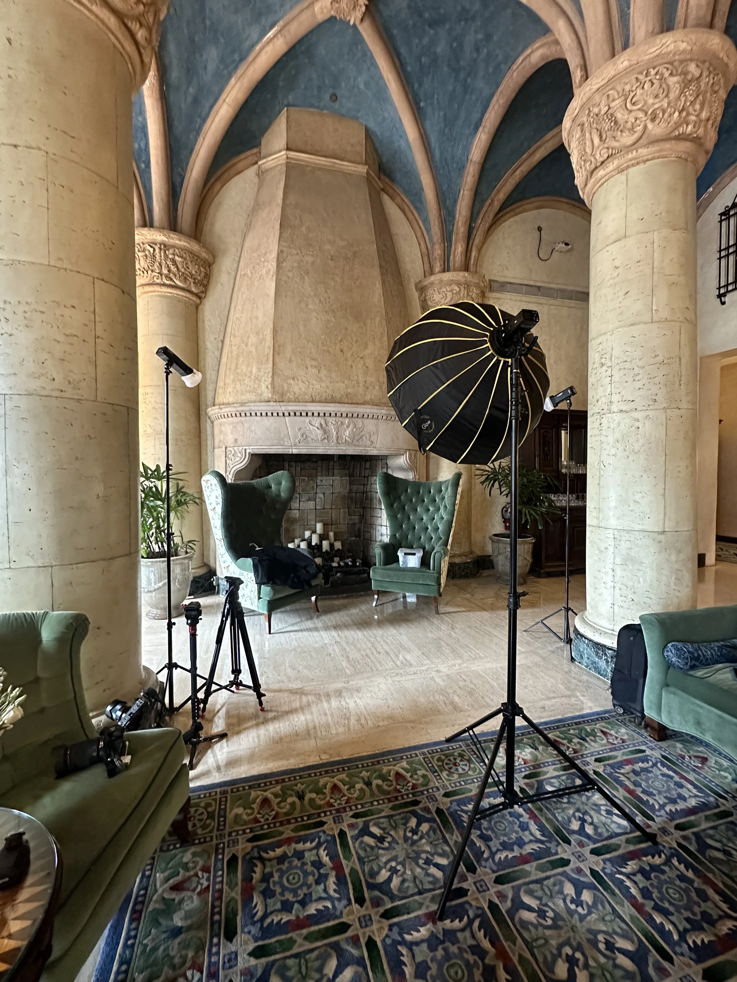 Interior of a luxurious room set up for a photo shoot with green chairs, photography equipment, and a large fireplace.