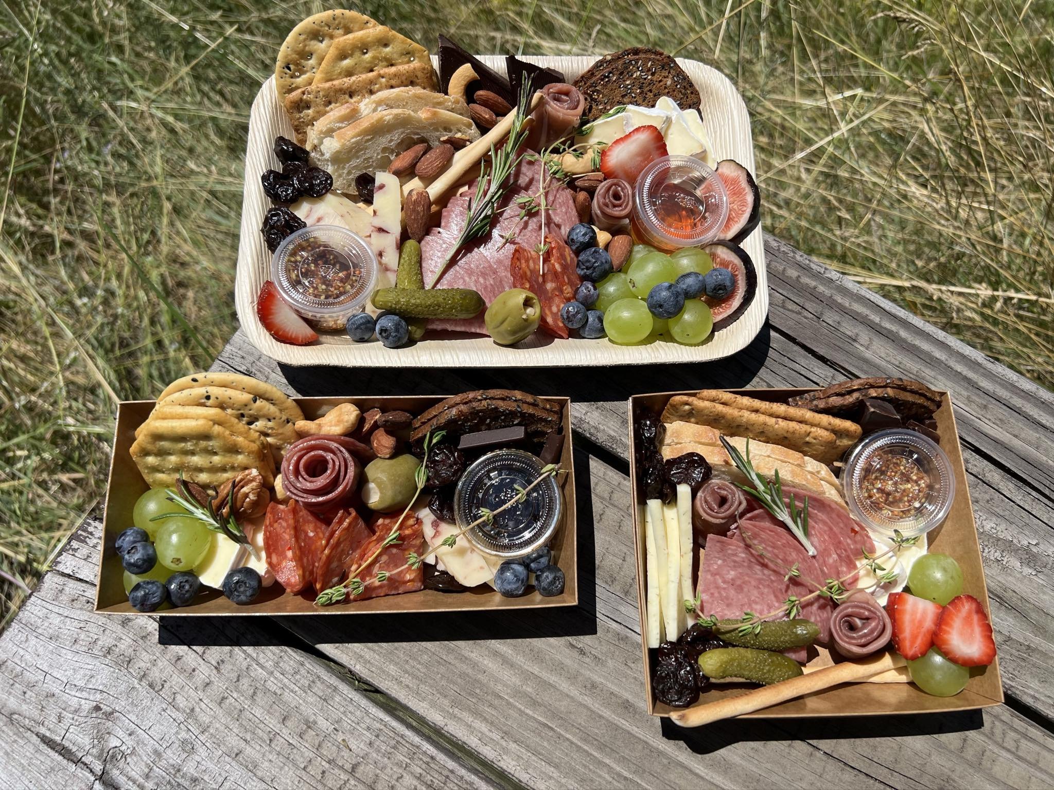 Three charcuterie boards containing assorted locally sourced meats, cheeses, crackers, fresh fruits like grapes and strawberries, almonds, pickles, and small containers of dips or spreads. Great for picnics, weddings, birthdays, and any special event