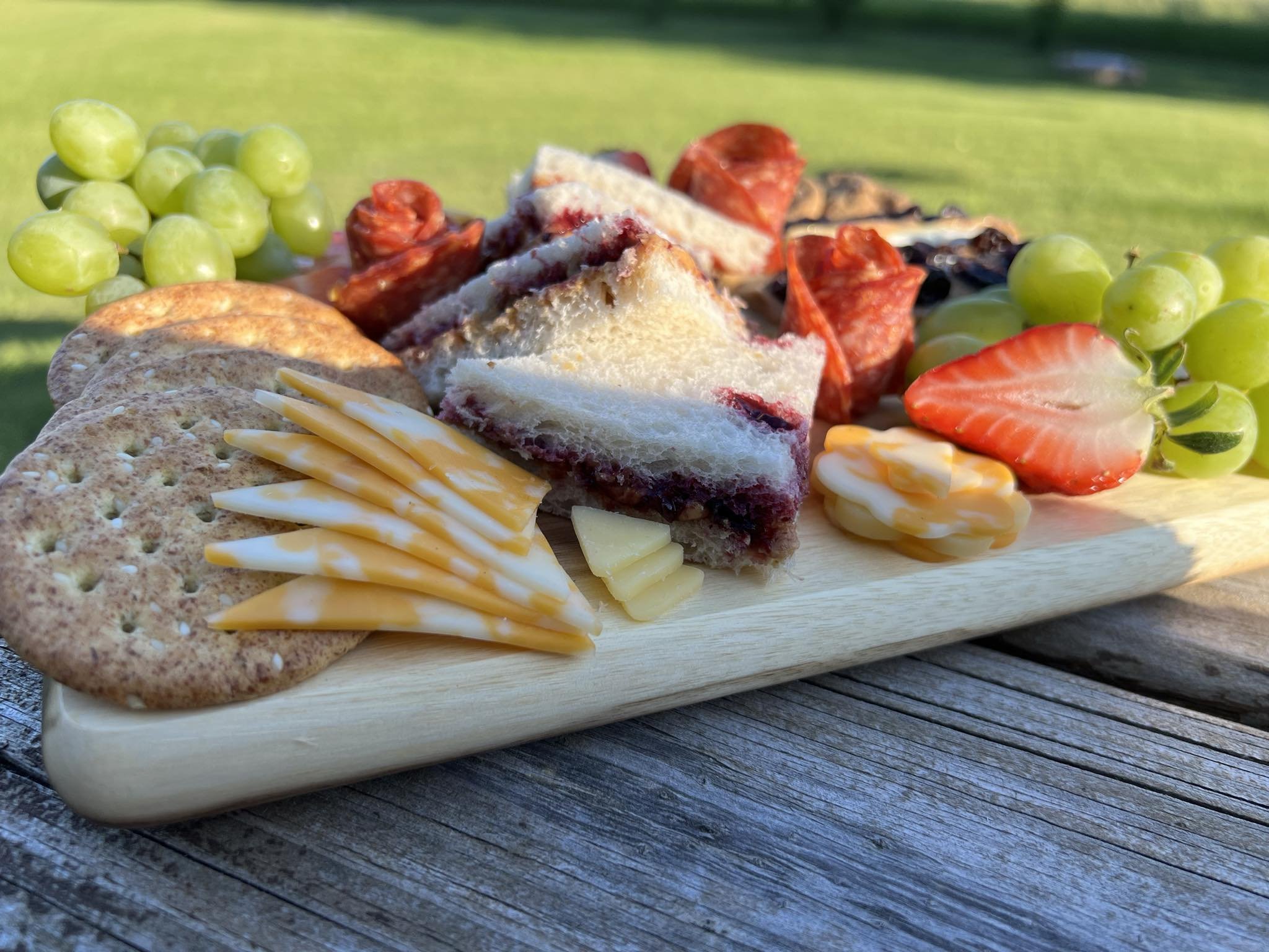 Charcuterie board with crackers, sliced cheese, green grapes, strawberry, sandwich triangles, and salami on wooden table outdoors.