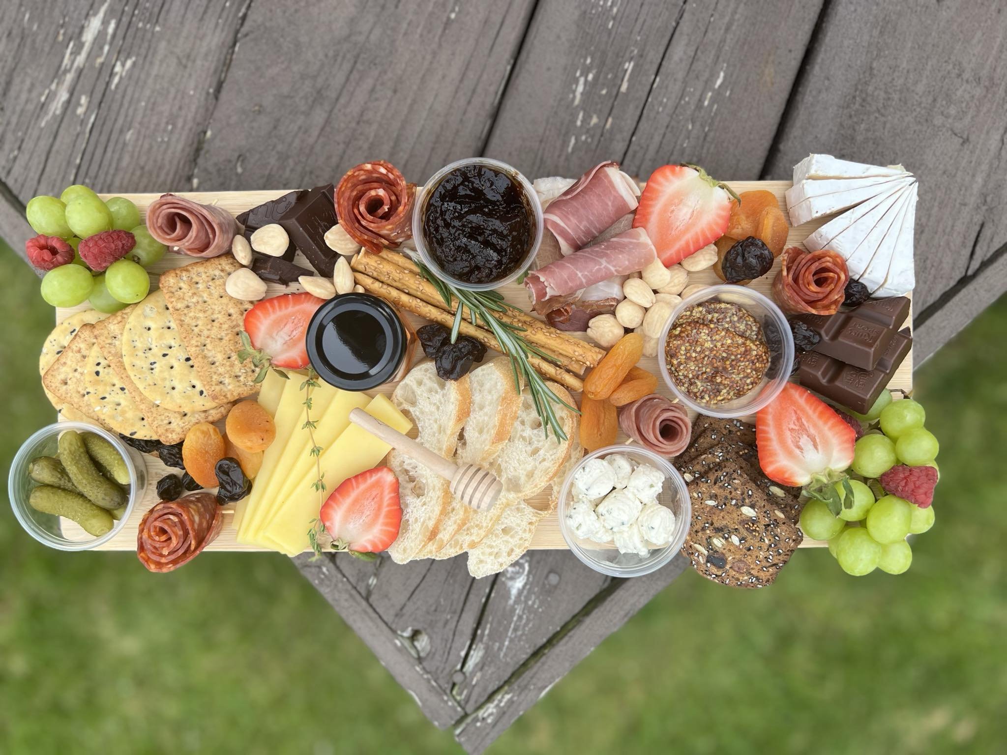 A charcuterie board on an outdoor table featuring assorted local cheeses, meats, bread, crackers, fruits, nuts, chocolate, pickles, and dips. Includes strawberries, grapes, apricots, almonds, and rosemary herb garnish.