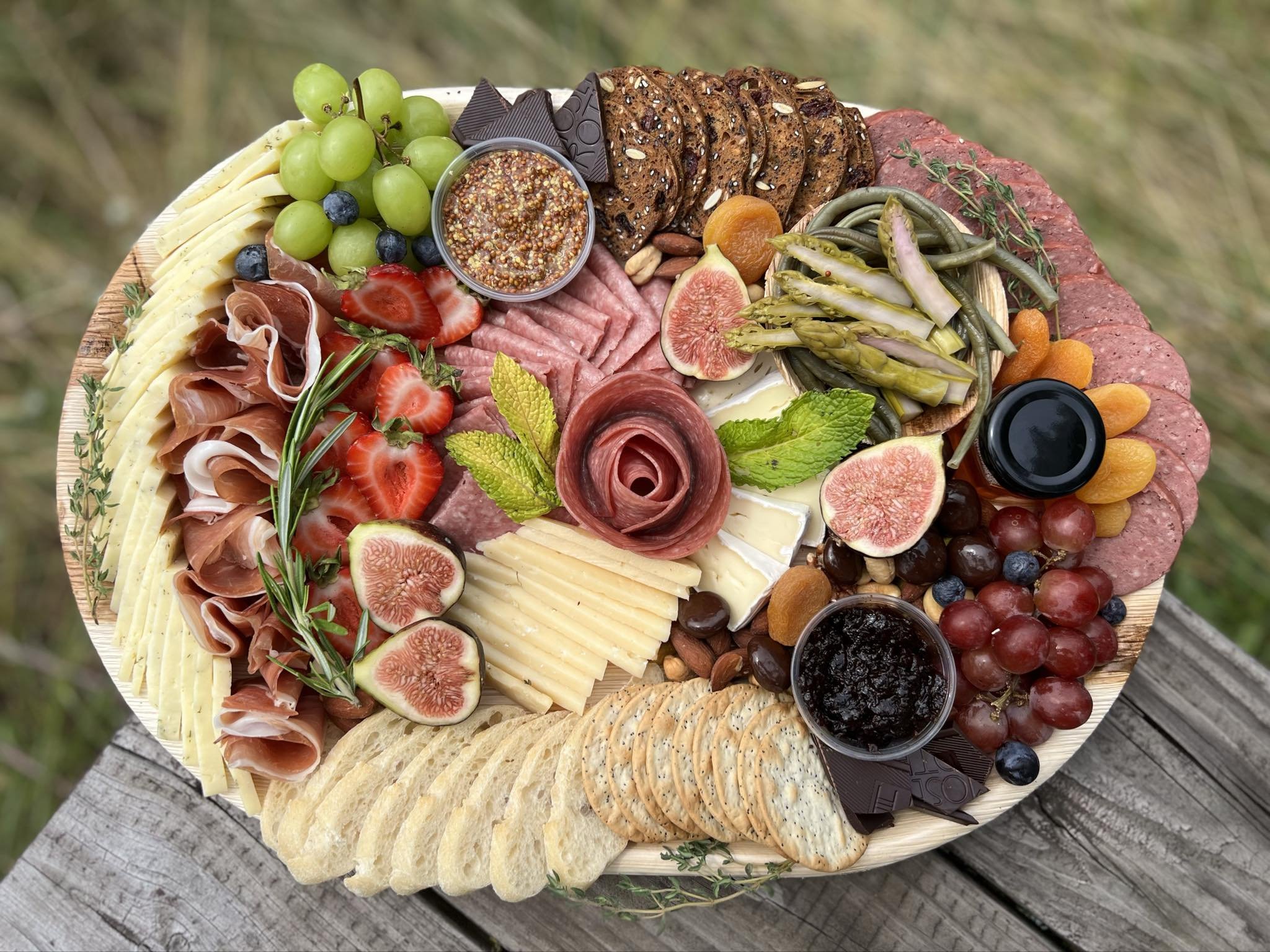 A charcuterie board featuring a variety of cheeses, cured meats, fresh fruits like grapes and figs, assorted nuts, crackers, and spreads, garnished with home garden grown herbs.