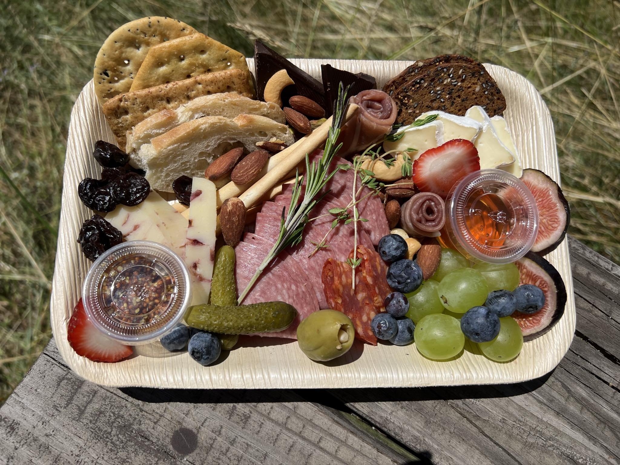 Charcuterie board with crackers, sliced bread, various cheeses, salami, prosciutto, olives, pickles, blueberries, grapes, figs, strawberries, almonds, cashews, dried fruit, rosemary, and small condiment cups on a wooden table.