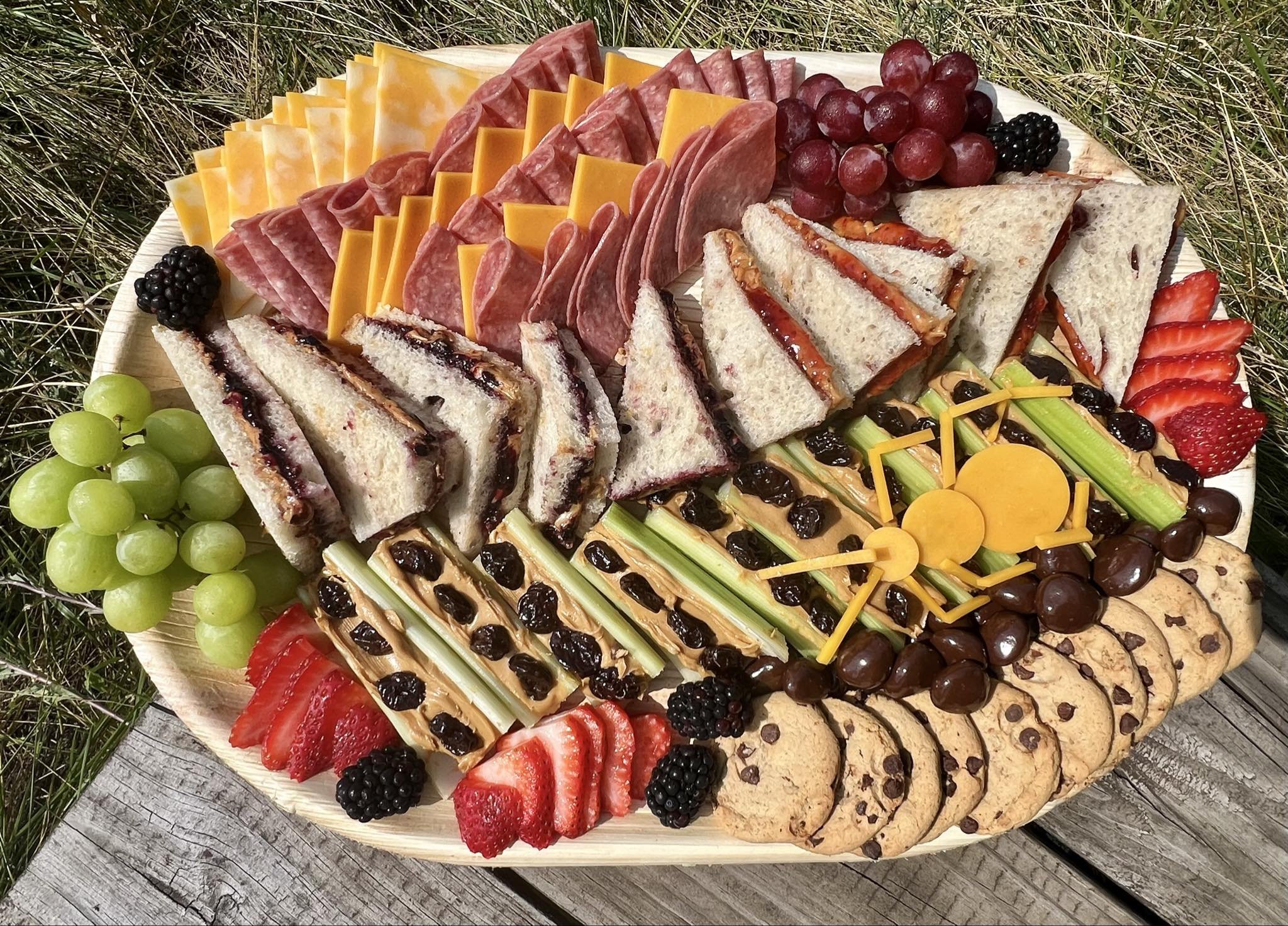 A kid's custom picnic charcuterie platter with assorted cheese slices, salami, PB&J sandwiches, ants on a log, grapes, strawberries, blackberries, and chocolate chip cookies! Yum! Order your custom themed charcuterie board today!