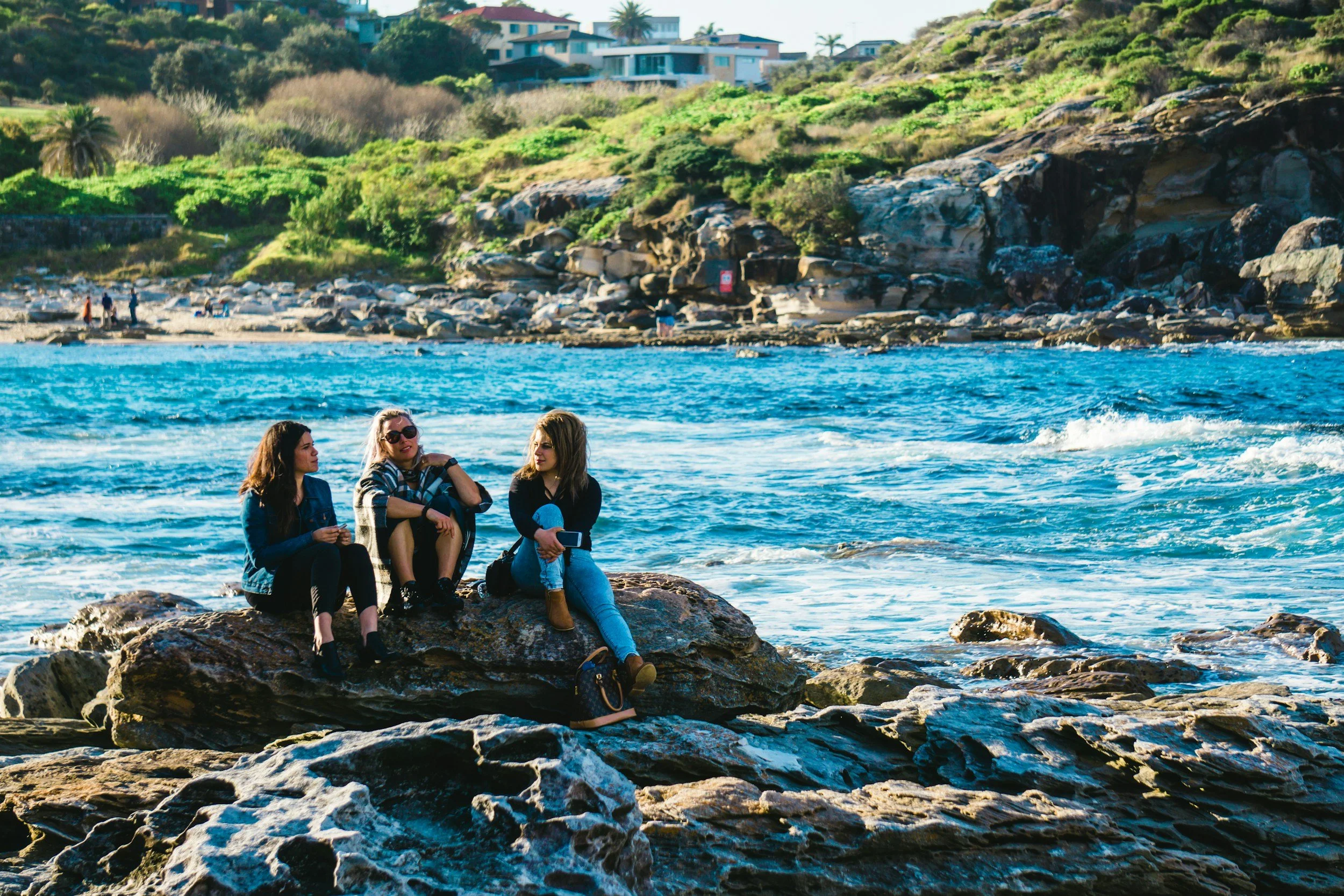 Three women sitting on rocks by the ocean shore, talking and relaxing with ocean waves crashing around them, in a sunny coastal area with houses and greenery in the background.