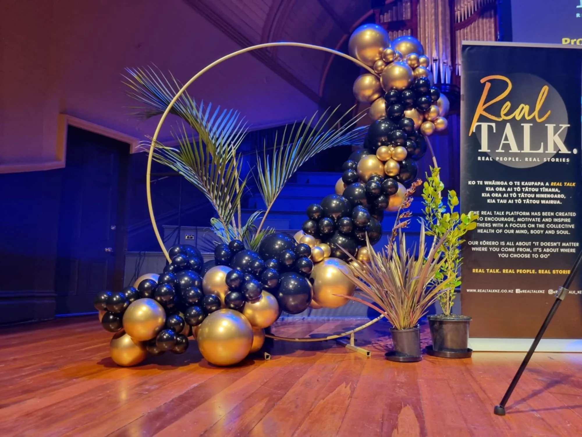 Decorative arrangement with black and gold balloons, palm leaves, and a vertical banner for 'Real Talk' event, displaying text about the platform's purpose and mission.