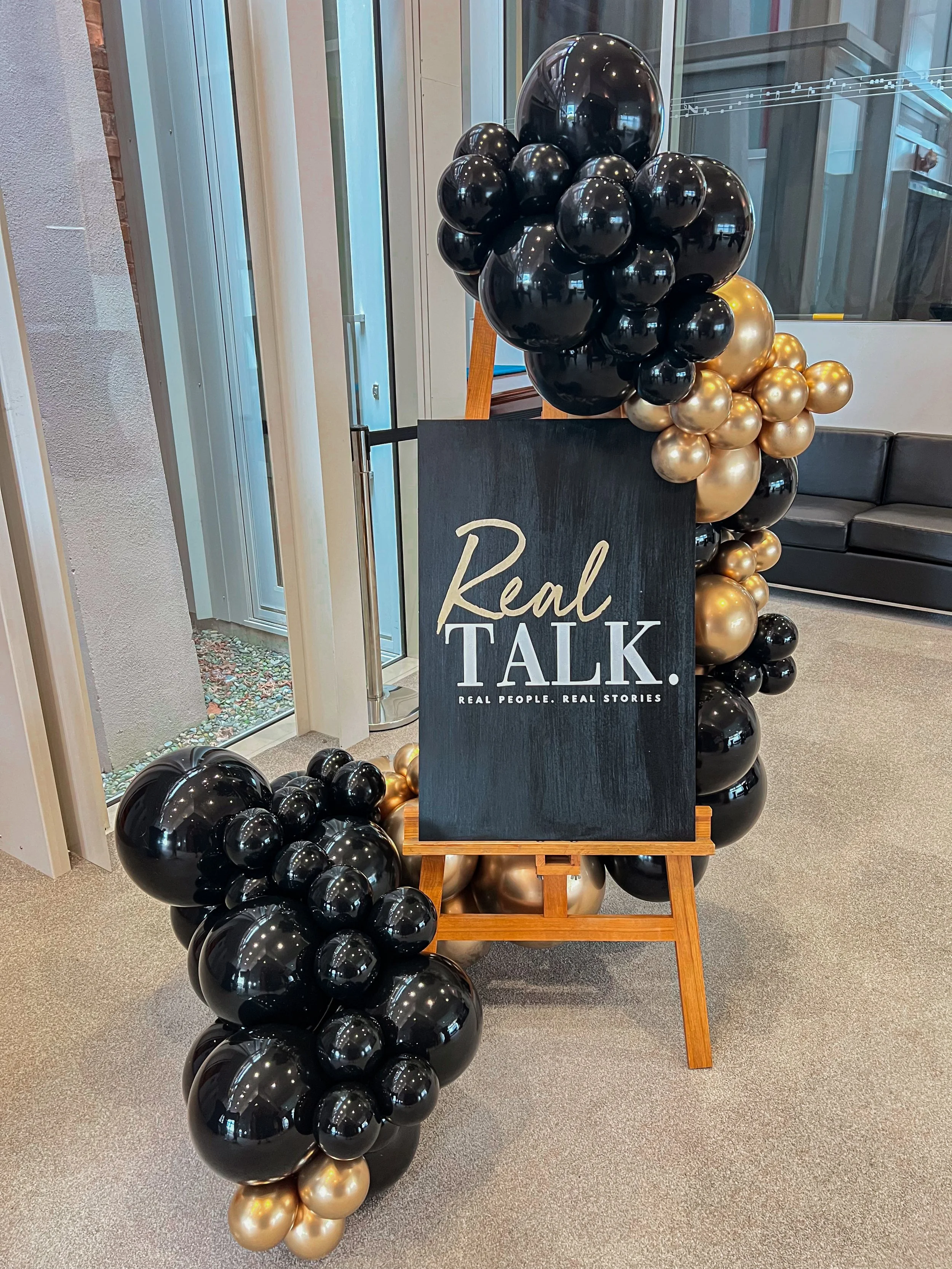 Black chalkboard sign with white and gold text saying 'Real TALK. Real People. Real Stories' surrounded by black, gold, and bronze balloons in an indoor setting.