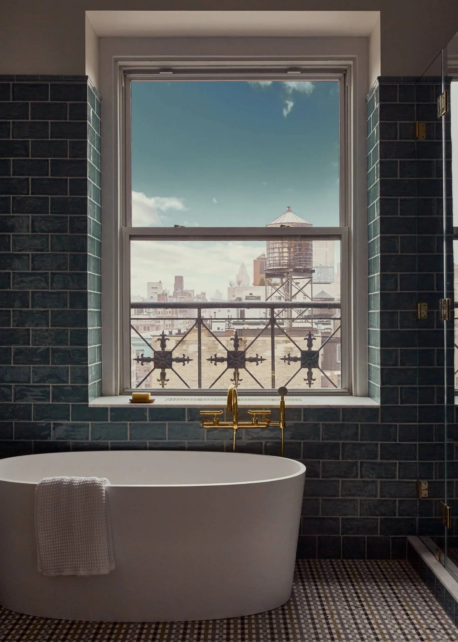 A white bathtub with a towel draped over the side, situated in a bathroom with dark tiled walls and a large window overlooking a cityscape with water towers and buildings.