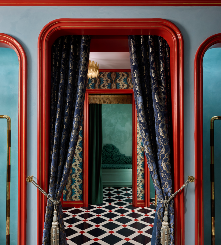 View through a richly decorated doorway into a room with ornate wallpaper, a bed with a patterned headboard, and curtains on the other side.