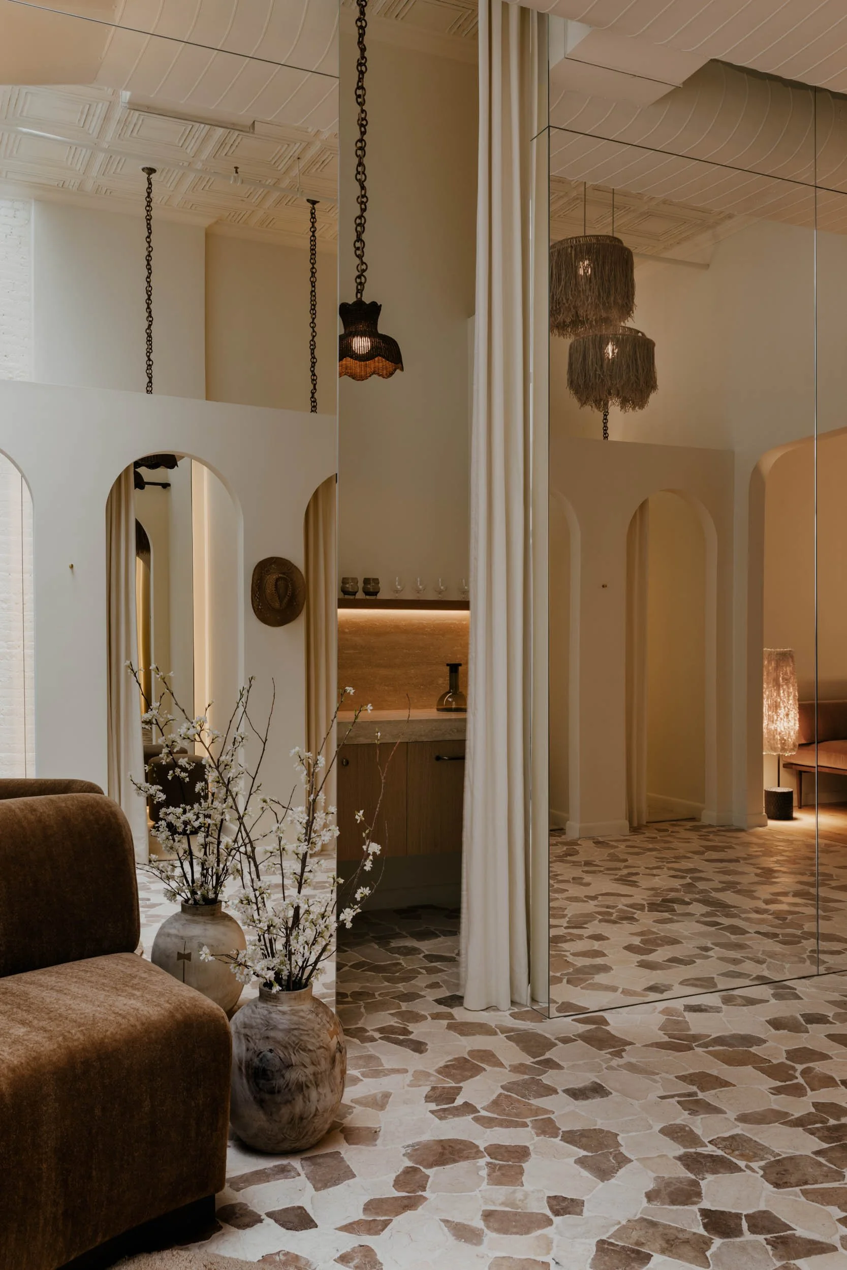 Interior of a room with beige walls and columns, decorative hanging lamps, a textured tile floor, and a mirror reflecting part of the room.