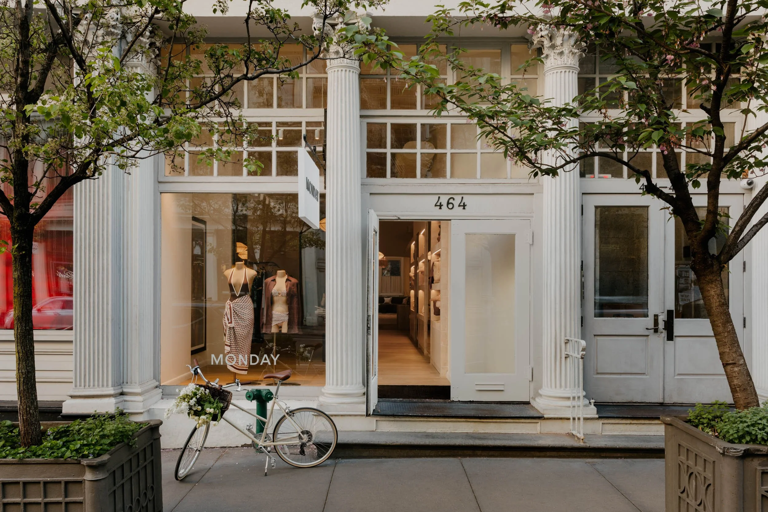 Storefront with large windows displaying mannequins dressed in clothing, flanked by white columns, with trees on either side and a bicycle parked in front.