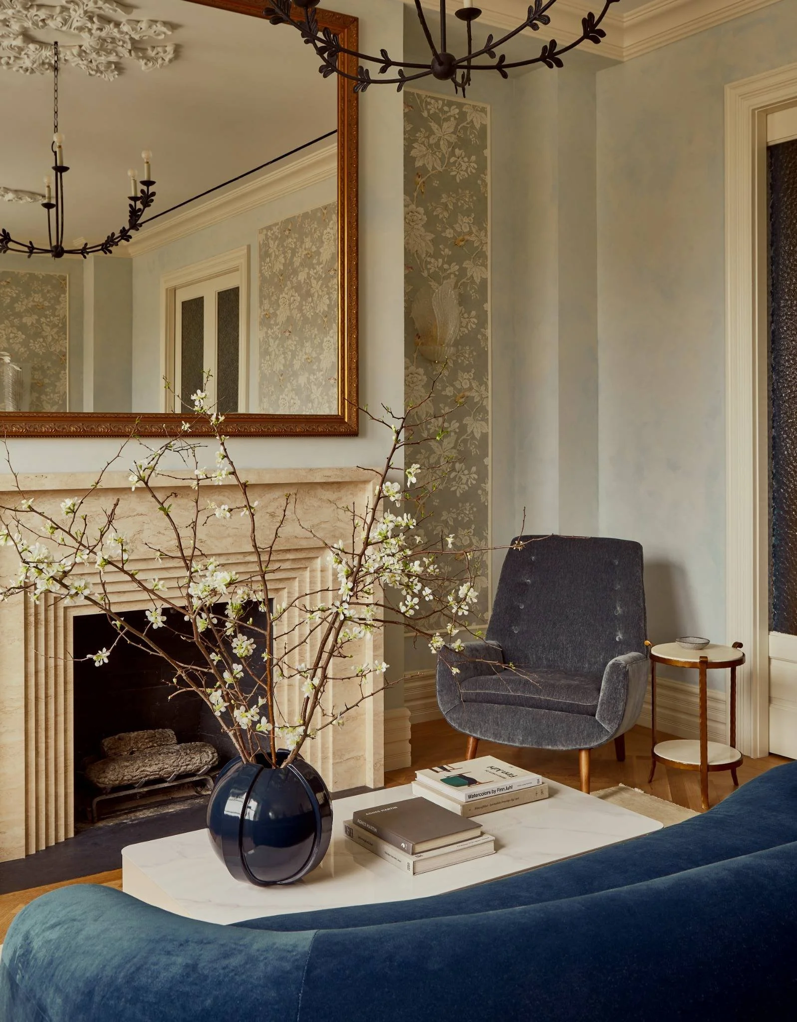 Living room with a marble fireplace, dark gray armchair, small side table, blue couch, books on a white coffee table, a large mirror above the fireplace, and a black decorative chandelier.