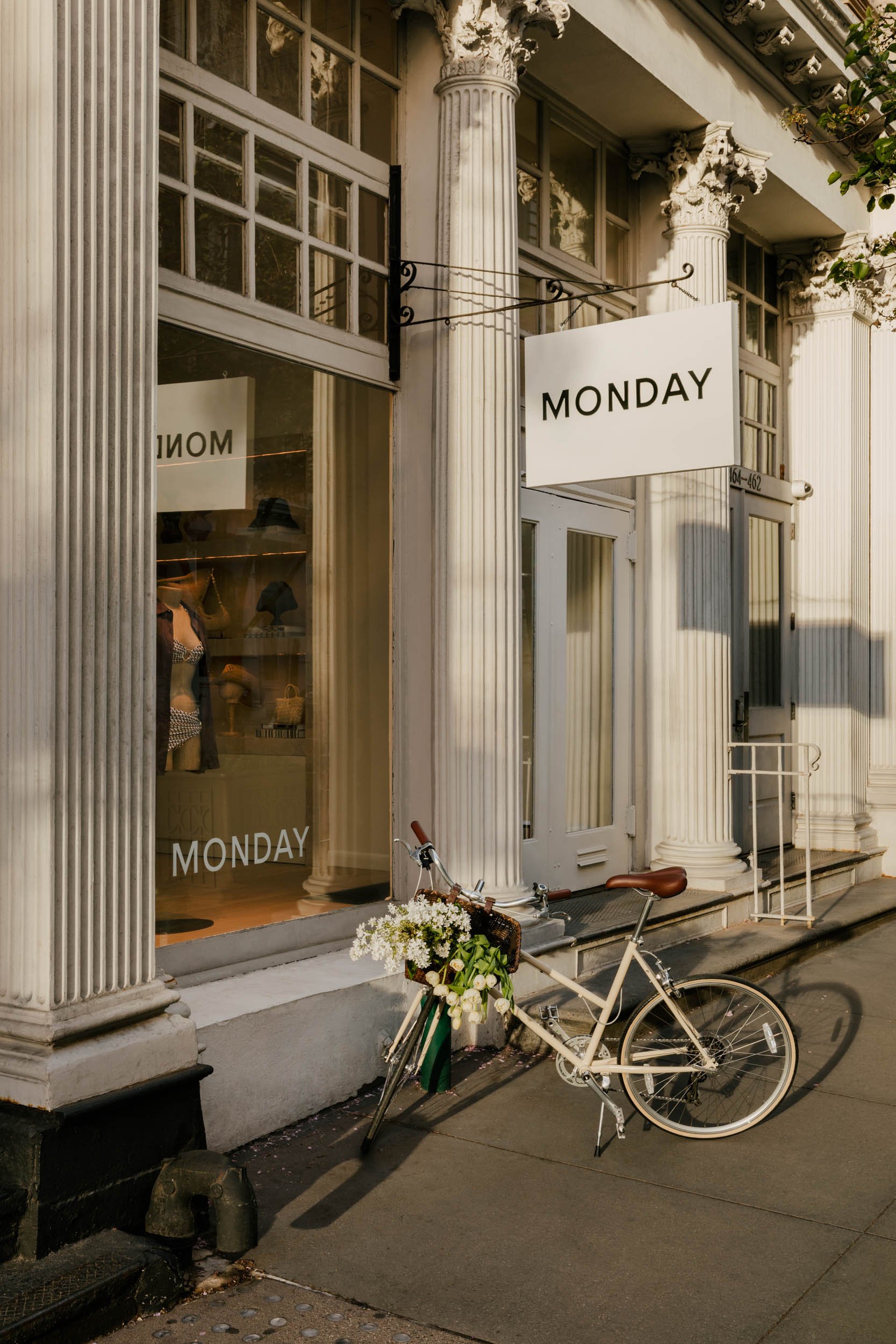 The image shows the exterior of a building with classical architectural design, featuring large white columns with ornate capitals. There is a hanging sign that says 'MONDAY'. A white bicycle with a basket full of white flowers leans against the building's steps, and a bicycle lock is visible on the ground nearby.