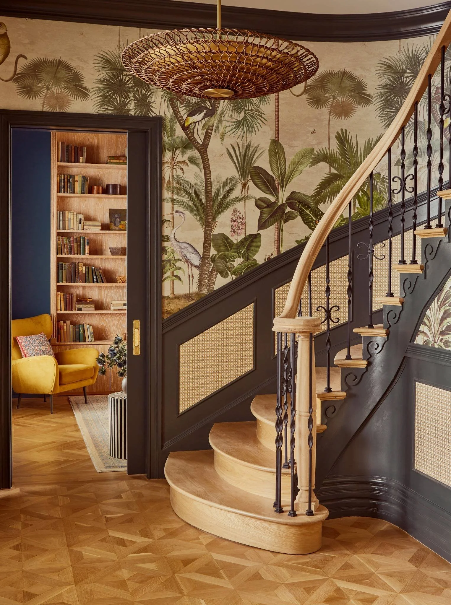 Interior of a house featuring a staircase with wooden steps and black iron balusters, a colorful wall mural of tropical plants and birds, and a room with a yellow armchair, bookshelves, and a decorative blanket.