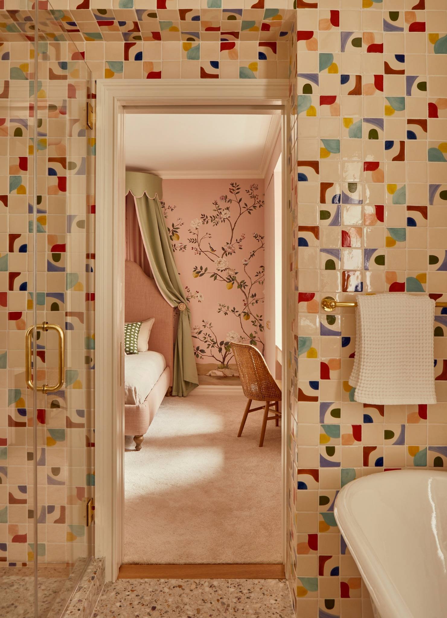 View from a bathroom with colorful mosaic tiles, showing an adjacent bedroom with floral wallpaper, a pink upholstered bed with a green curtain, and a wicker chair.