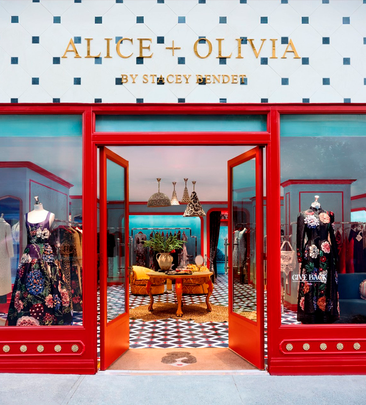 Storefront with red framing and open doors, signage reads "ALICE + OLIVIA BY STACEY BENDET," showing a boutique with dresses and mannequins, a patterned floor, and interior decorations including a table with a plant and seating.