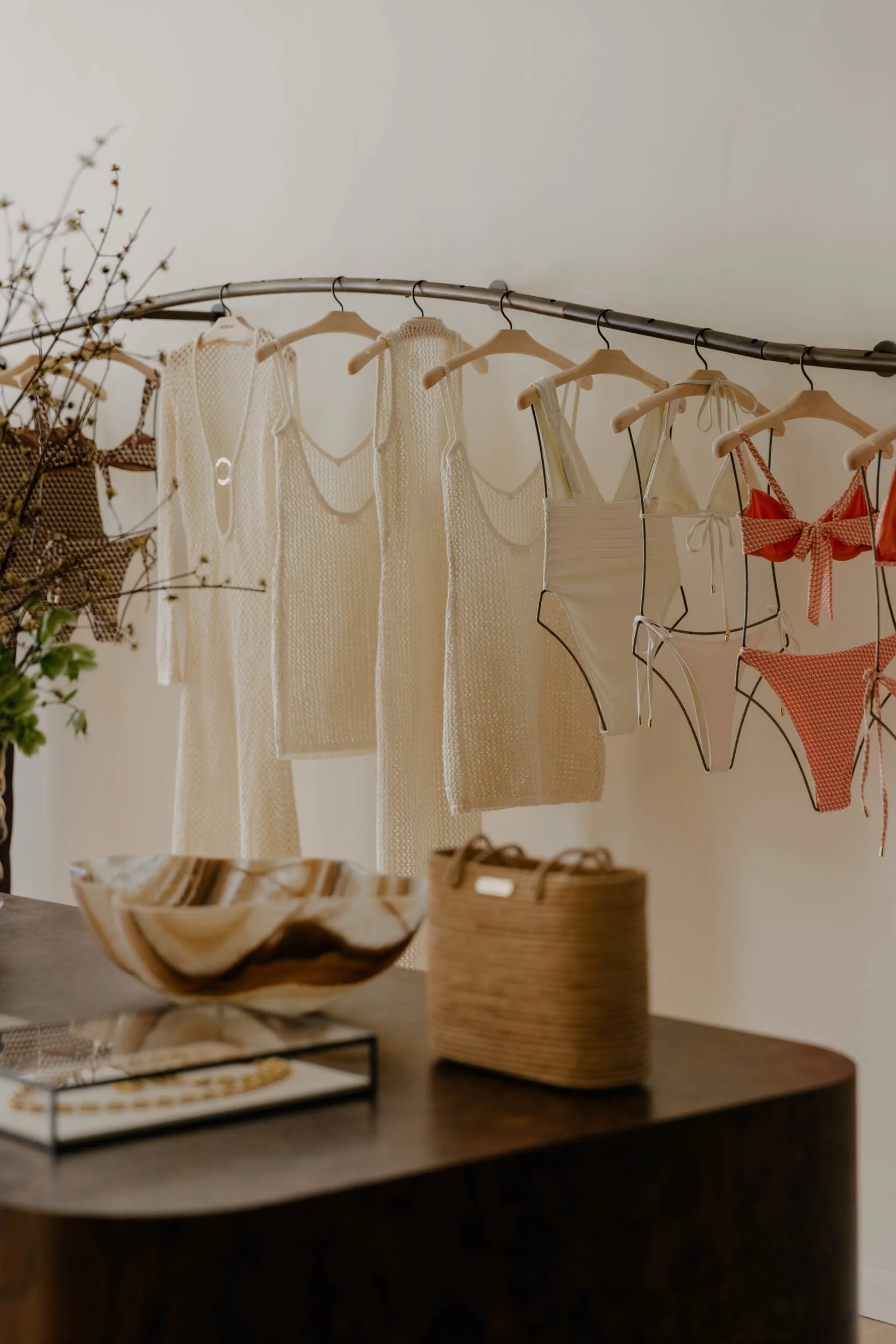 Clothing items such as dresses and swimsuits hanging on a curved metal rack in a boutique or store, with a wooden table displaying woven baskets and other decorative items in the foreground.