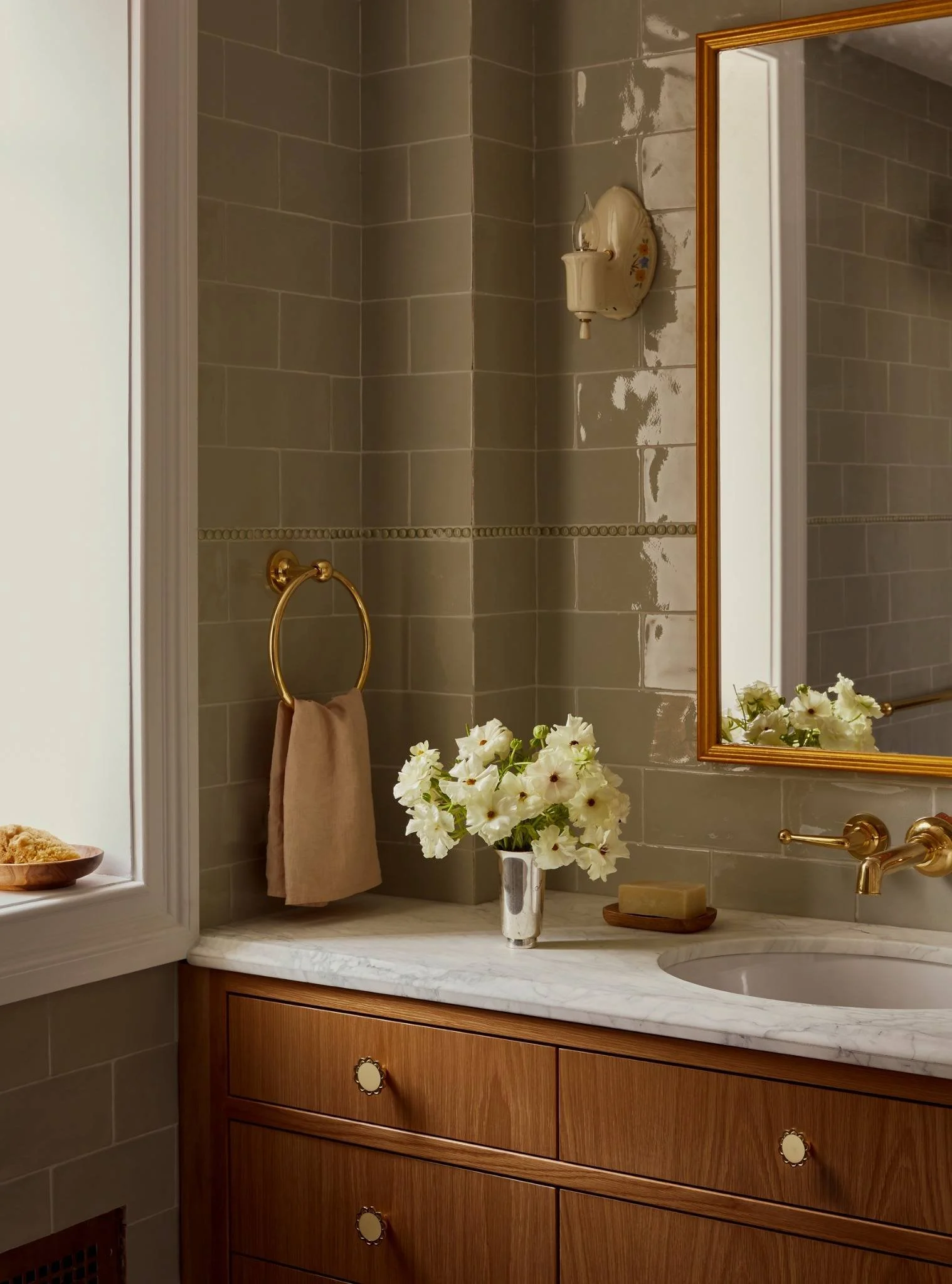 Bathroom with beige tiled walls, a wooden vanity with a marble countertop, a vase of white flowers, a bar of soap, a gold towel ring with a towel, a wall-mounted light fixture, and a mirror with a gold frame.
