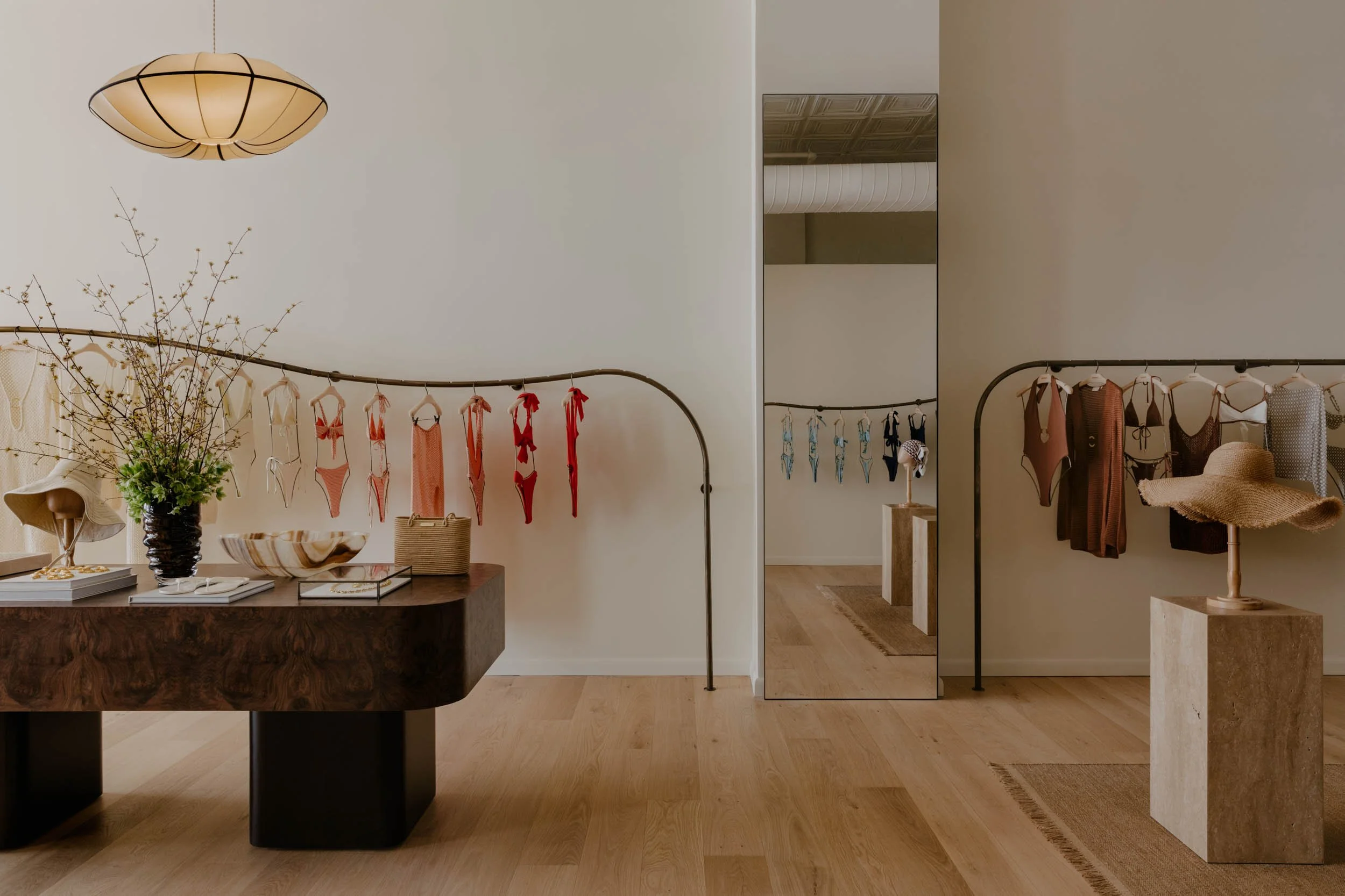 Interior of a boutique store displaying swimsuits hanging on black metal racks, with a large mirror on the right and a wooden table with decorative items and jewelry, beige walls, and wooden flooring.