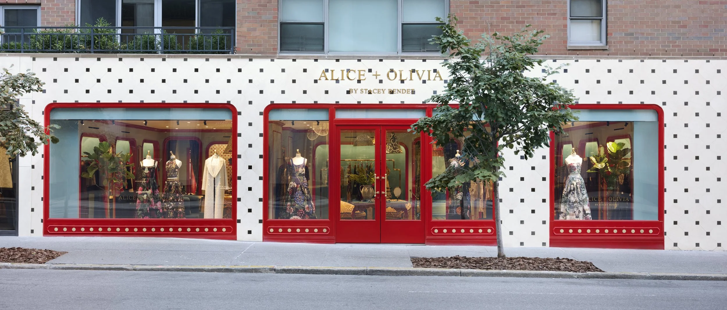 Front view of clothing boutique with large display windows and red framing, showcasing mannequins dressed in floral dresses, located on a city street with a sidewalk, small tree, and a brick building in the background.