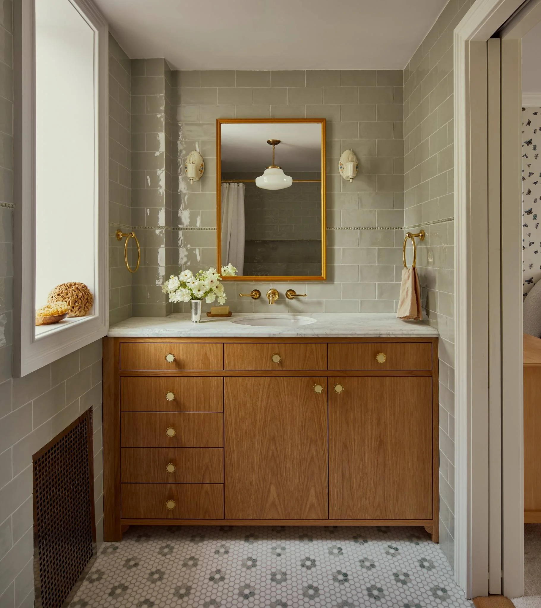 Bathroom vanity with a wooden cabinet, a white marble countertop, a mirror, and gold fixtures, including a soap dish, towel rings, and faucet, with a small flower arrangement and sponge on the countertop.