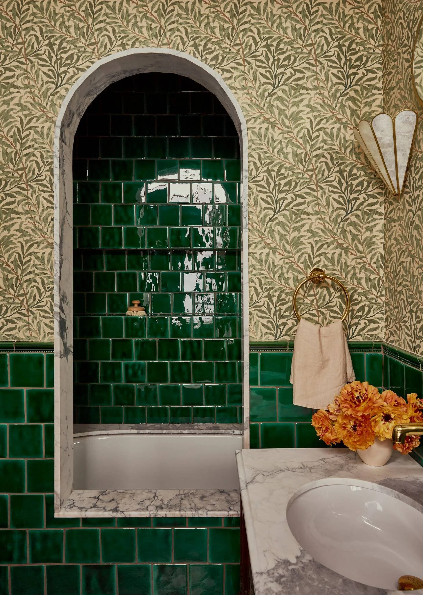 A bathroom with green tiled shower stall, marble accents, floral wallpaper, gold fixtures, a towel ring with a beige towel, and a marble countertop with a vase of orange flowers.