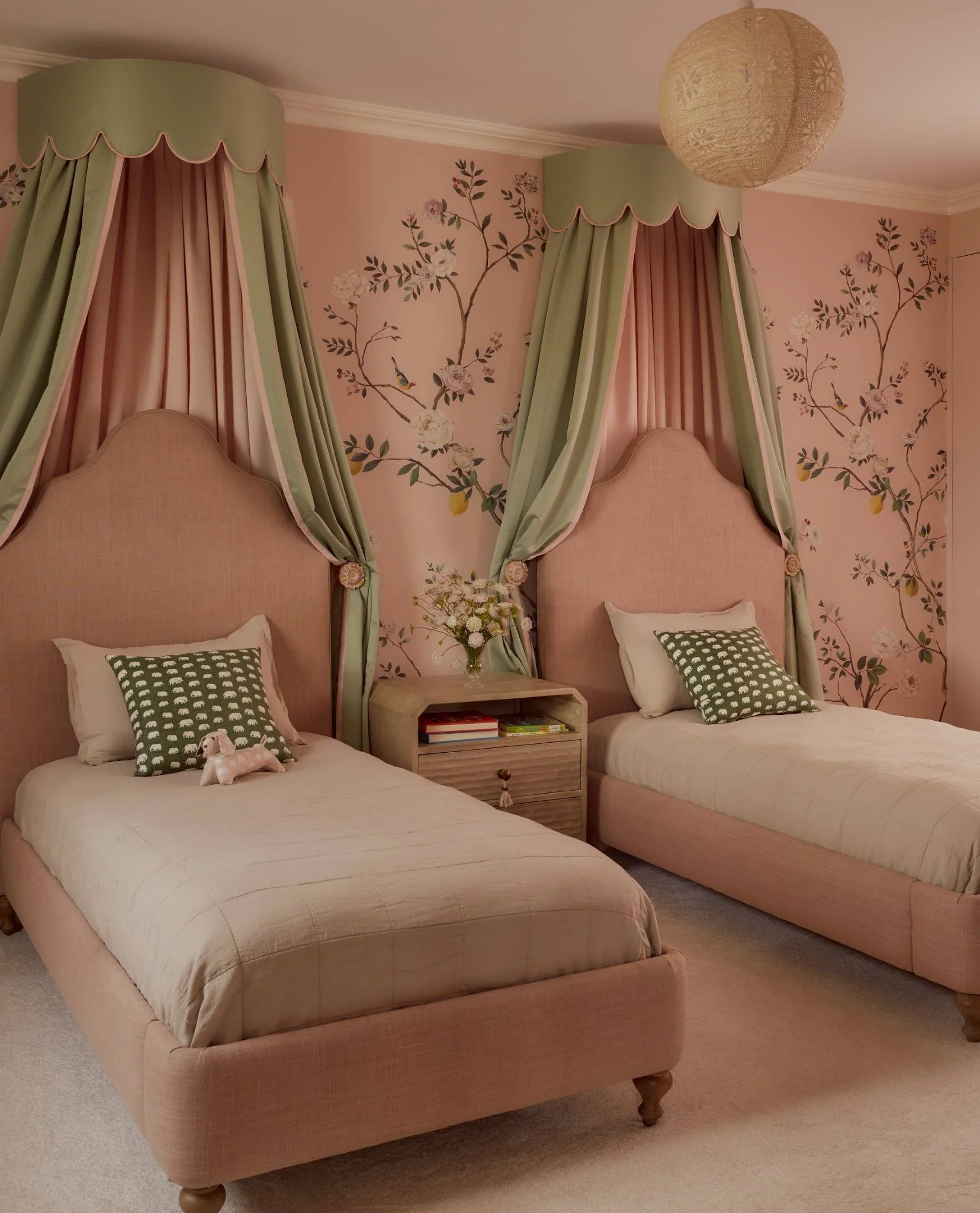 A bedroom with two twin beds featuring pink headboards, green and pink draped curtains with floral canopy over each bed, floral wallpaper with birds, a small bedside table with books and a vase of flowers, and plush toys on the beds.