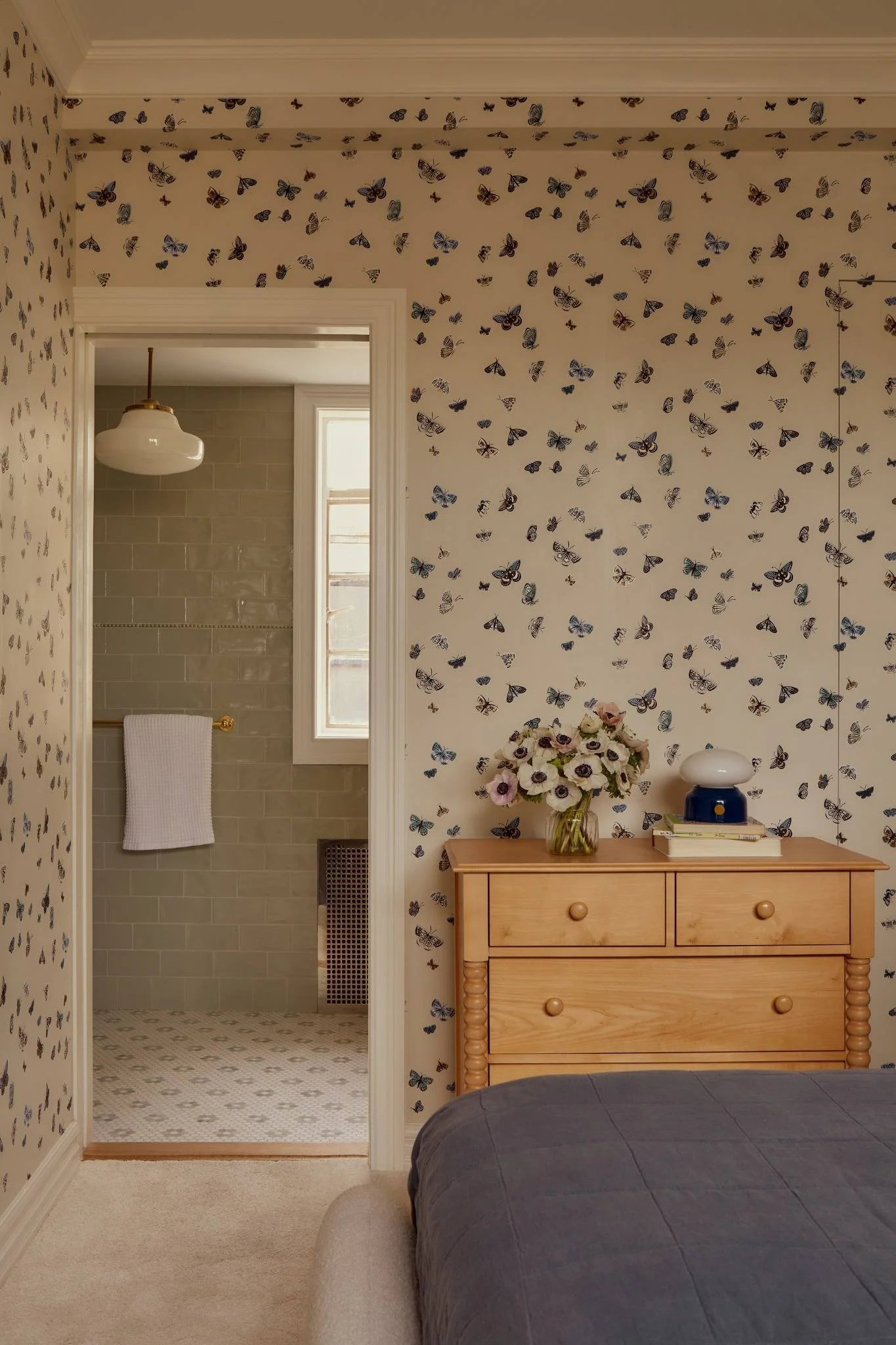 A bedroom with wallpaper featuring blue and black butterfly patterns, a wooden dresser with a vase of flowers and a lamp, and a view into a bathroom with beige subway tiles and a white towel hanging on a gold bar.