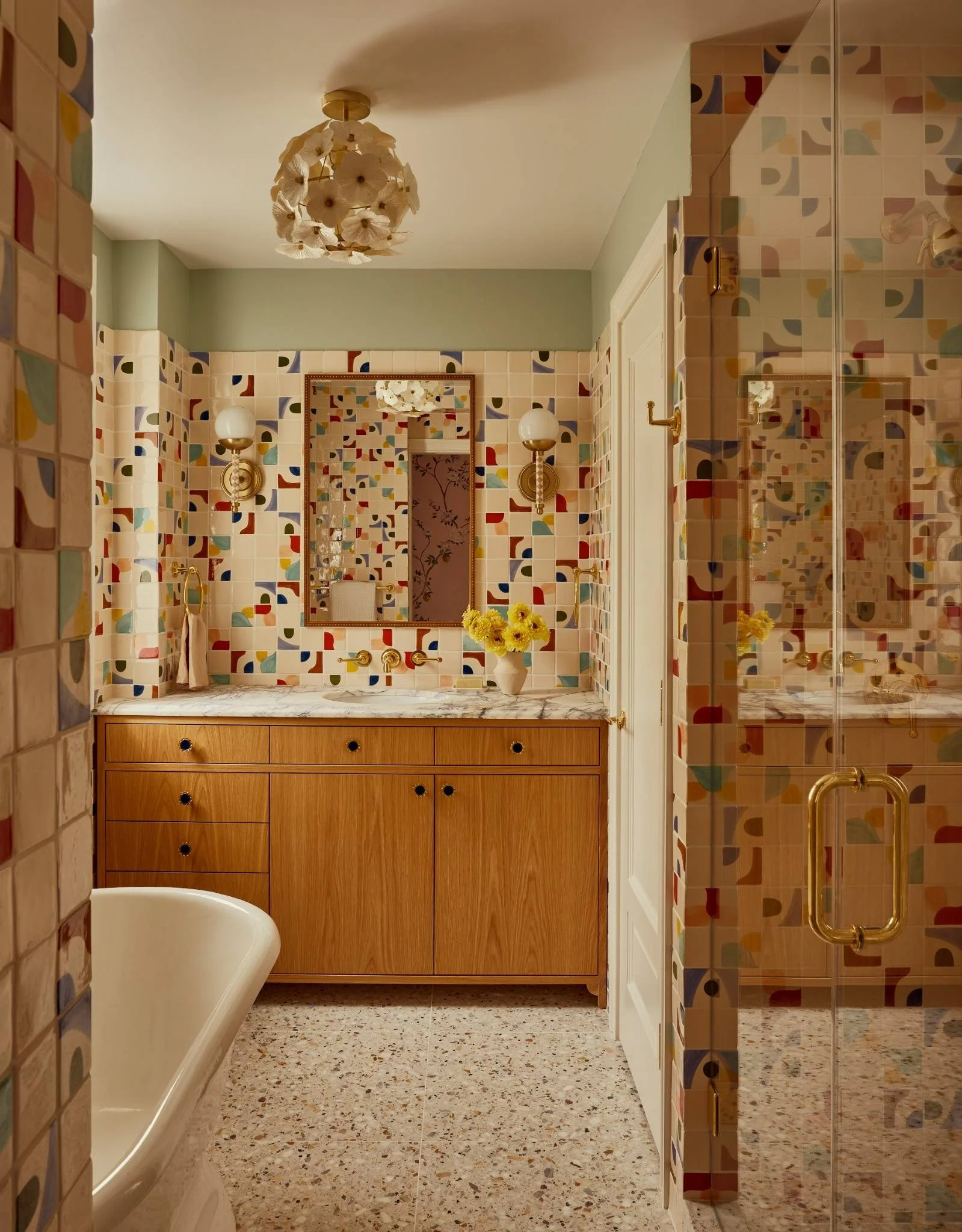A bathroom with colorful patterned tiles on the walls, a wooden vanity with a marble countertop, a mirror, and a vase with yellow flowers, a bathtub, and a glass shower enclosure with gold hardware.